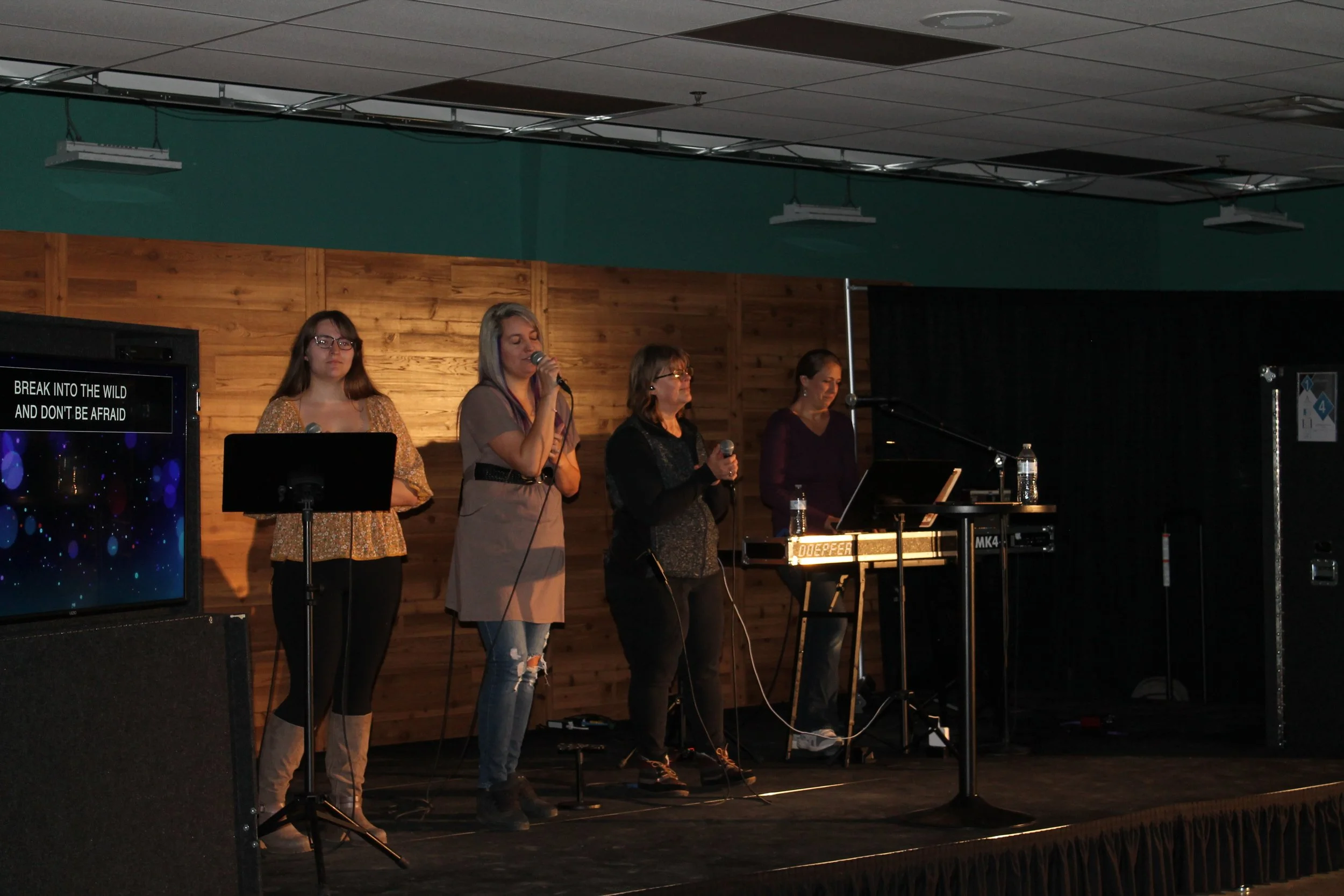 Four women singing into microphones on stage, with a keyboard and water bottles, in front of a wooden wall in a dimly lit room.