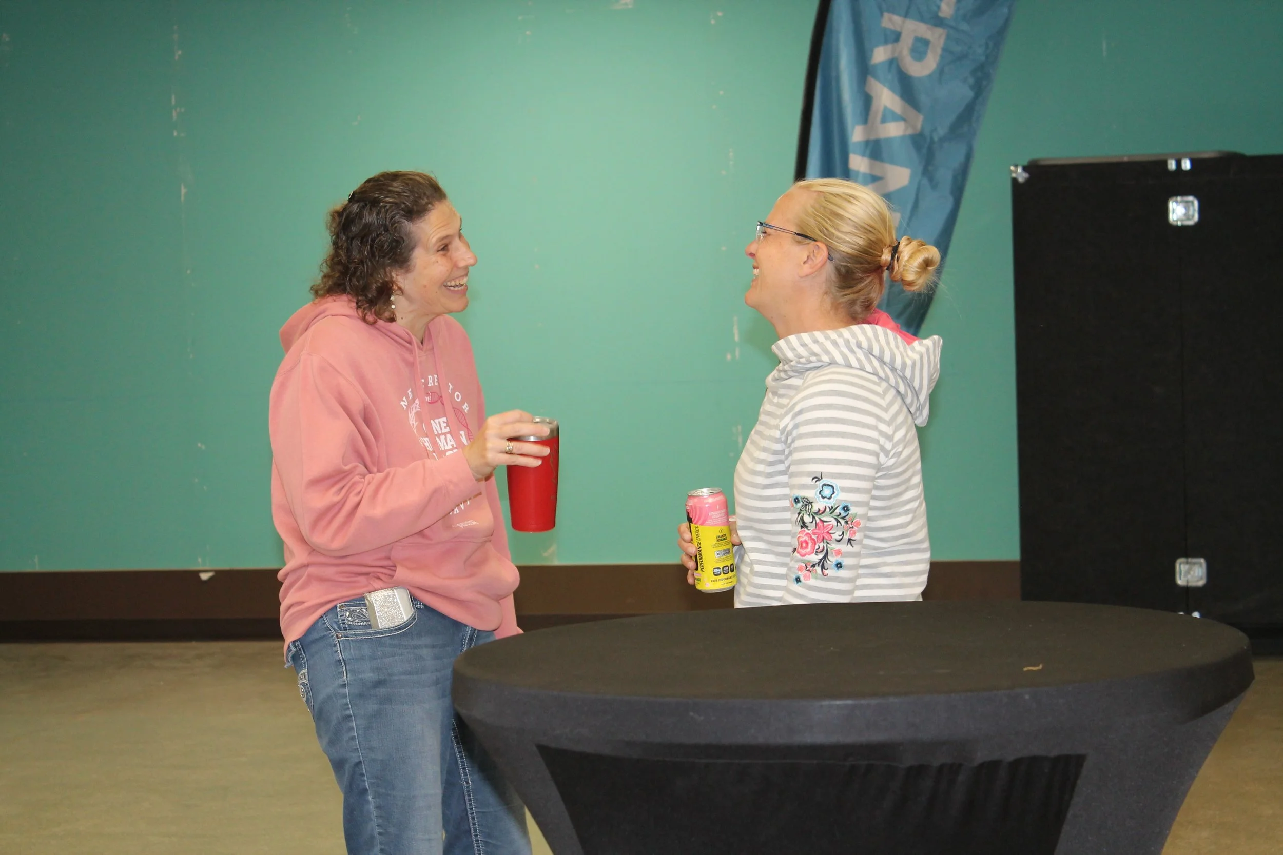 Two women smiling and talking while holding drinks, standing near a round black table, with a teal wall and a banner in the background.