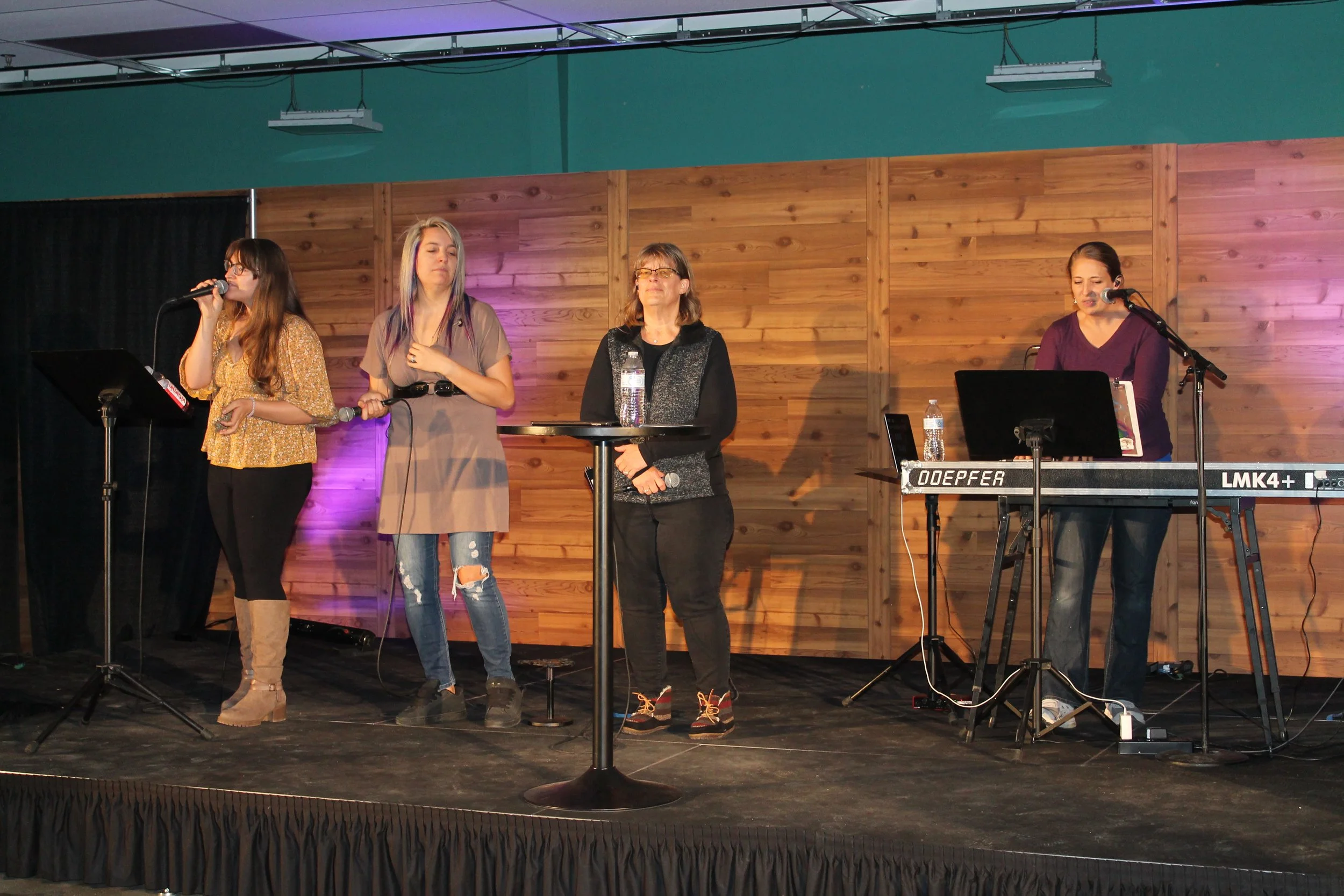 Four women on stage, three with microphones and one at a keyboard, performing in front of wooden panel backdrop with purple lighting.