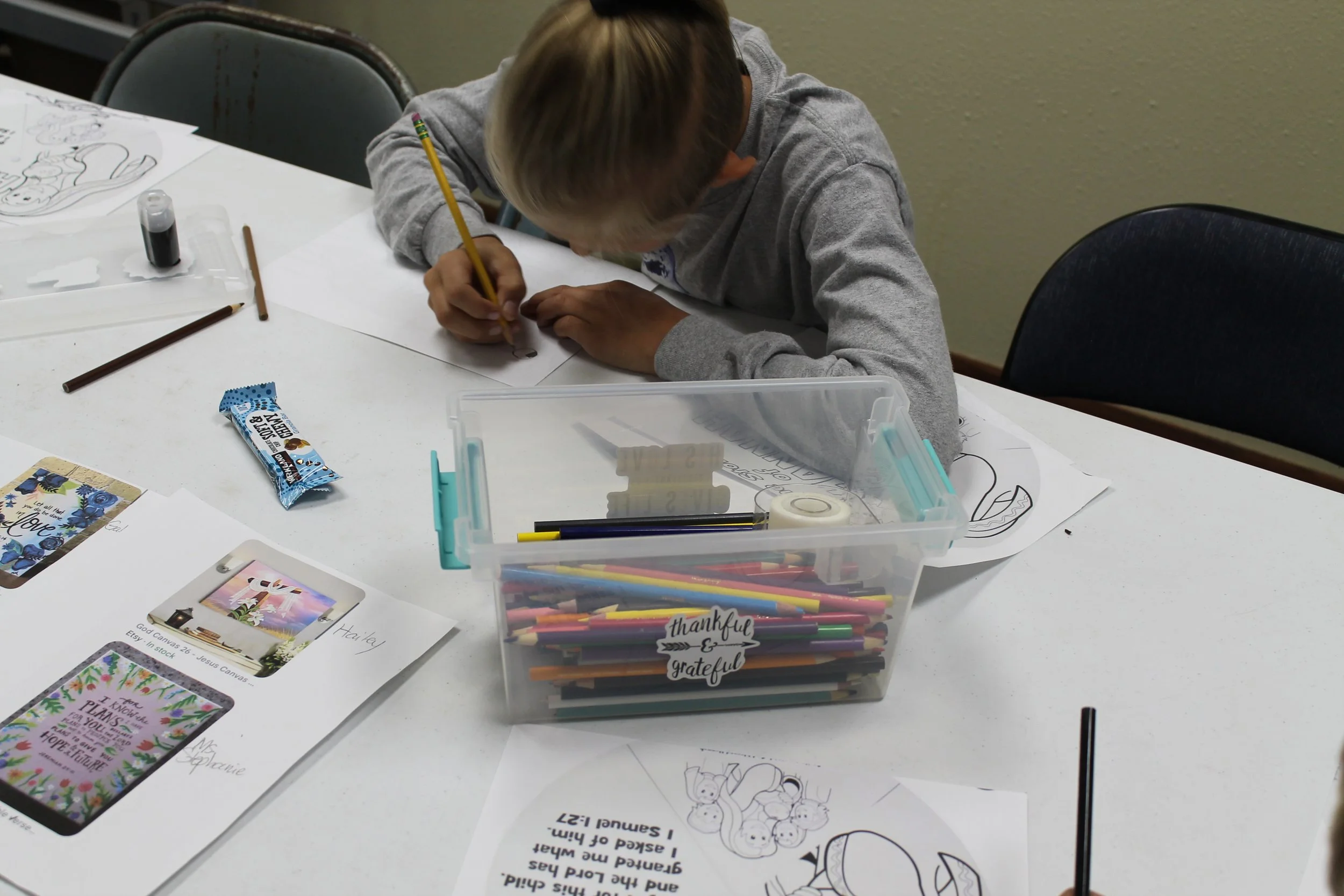A young child with blond hair wearing a gray hoodie is sitting at a white table, coloring on paper with a yellow pencil. The table has various coloring pages, a clear plastic box filled with colored pencils, markers, and other art supplies, and a sma