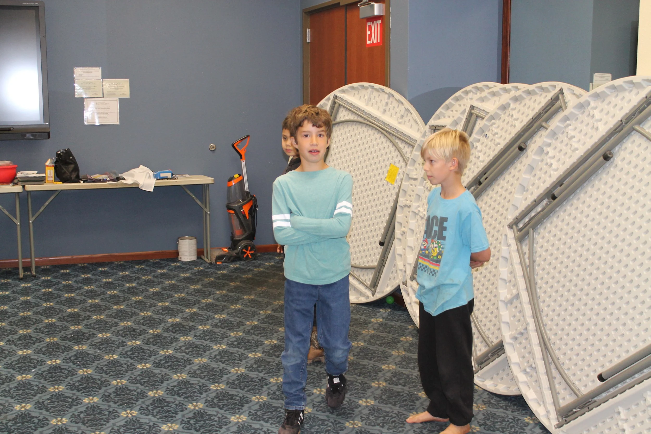 Three children in a room with folding tables and chairs, some folded tables leaned against the wall, and an exit door in the background.