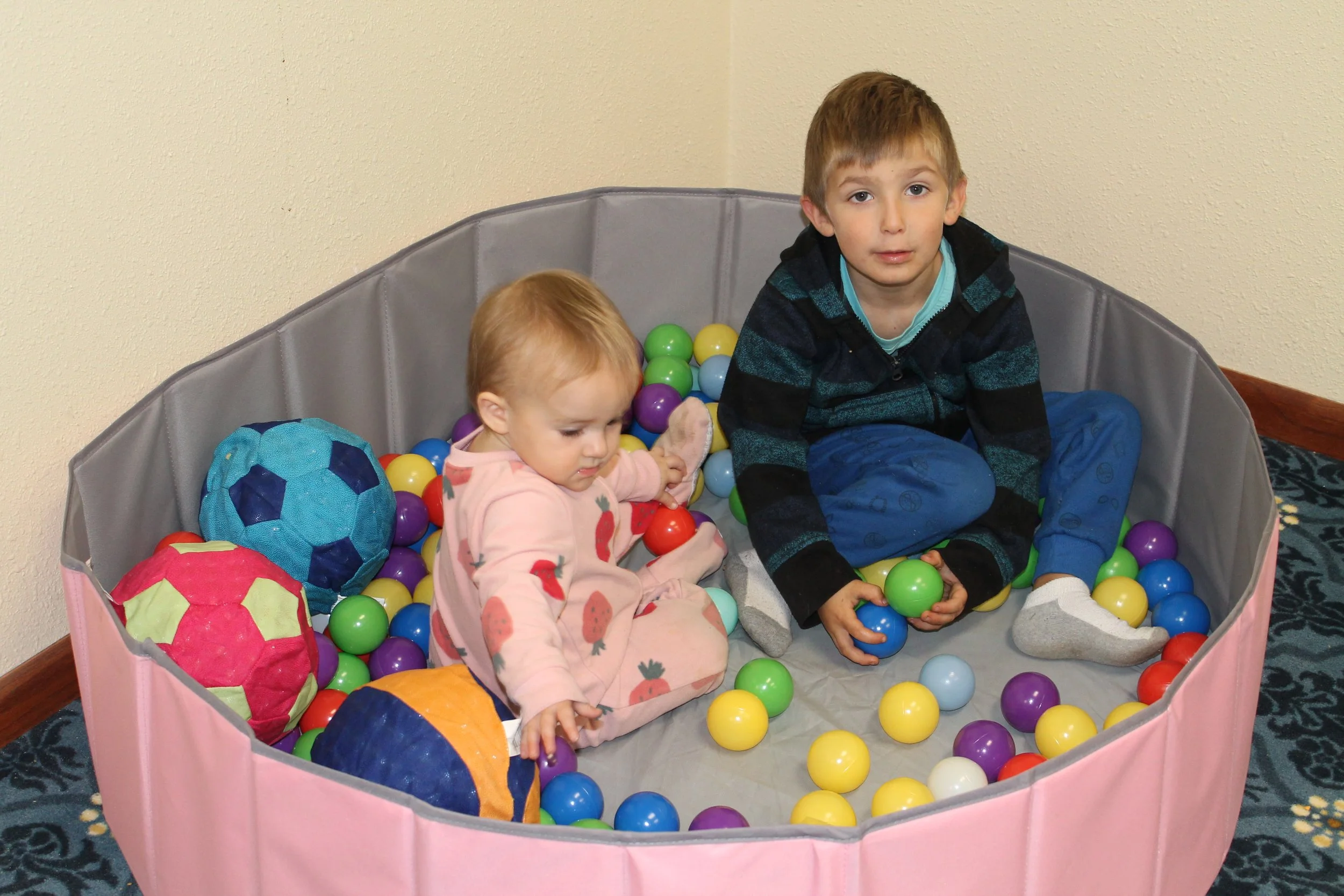 Two children, a young girl and an older boy, sitting in a ball pit filled with colorful plastic balls. The girl is wearing pink pajamas with strawberry prints, and the boy is wearing a dark hoodie with blue pants. They are playing with the balls inside the ball pit, which is pink with a grey interior, located in a room with beige walls and a patterned carpet.