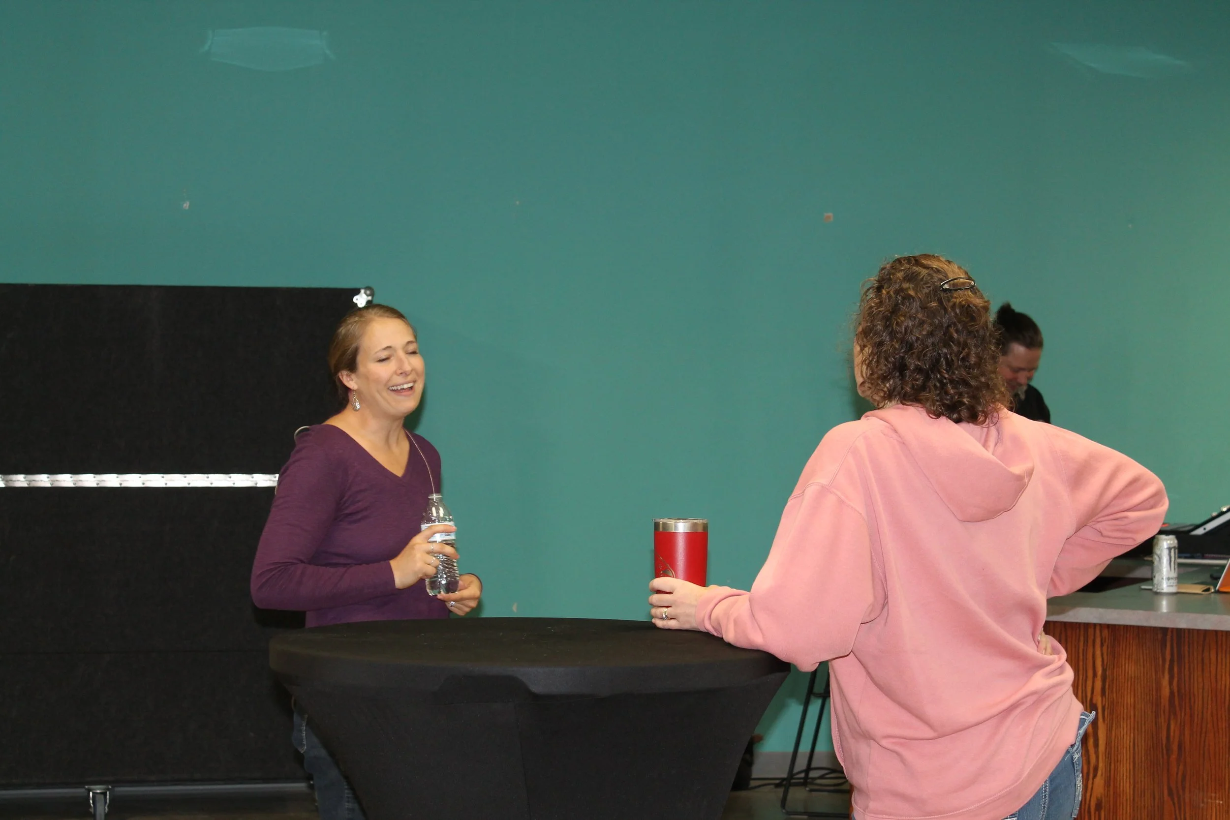 Two women with curly hair are standing at a black table, smiling and engaging in conversation. One woman is holding a water bottle, and the other is holding a red tumbler.