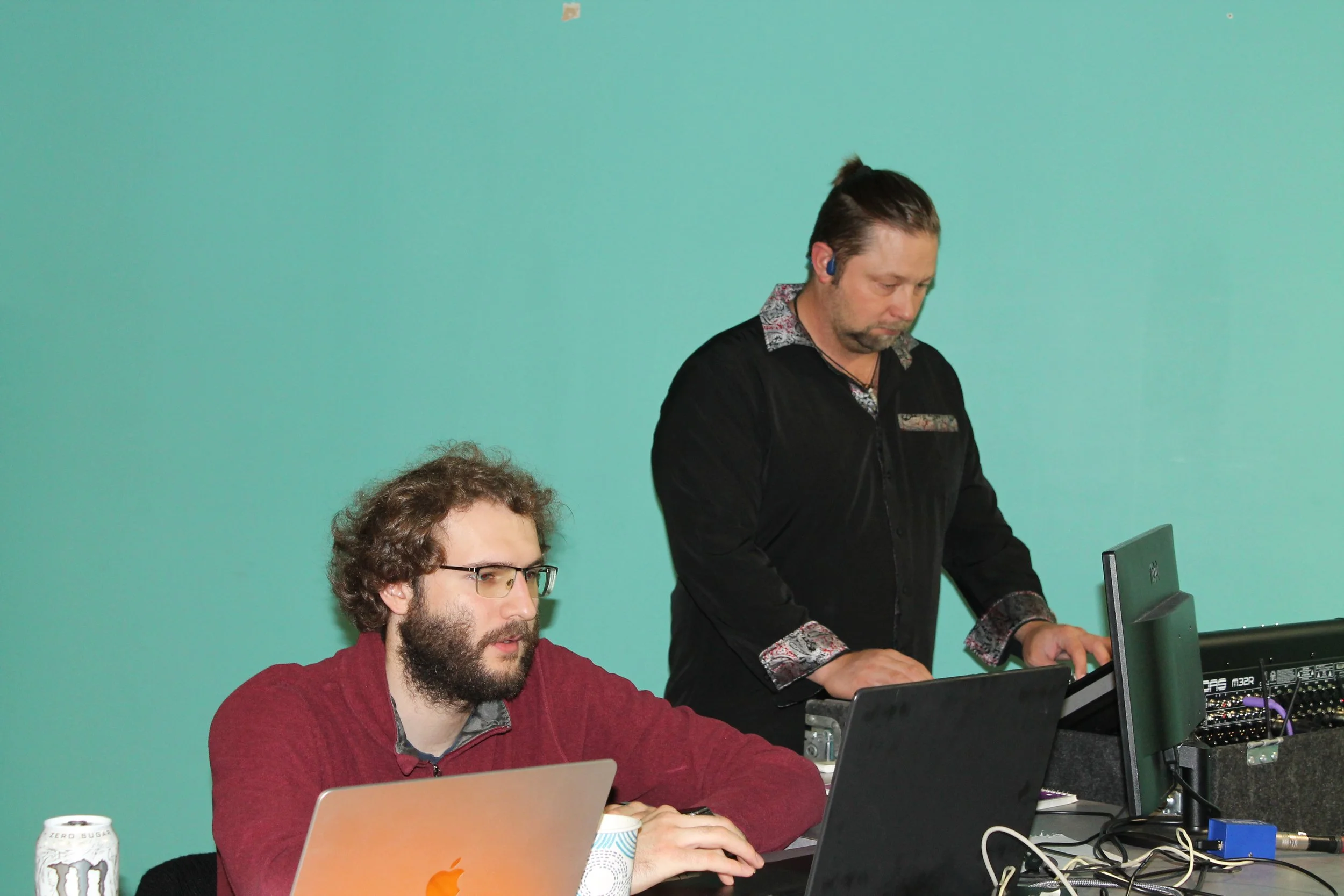 Two men working at a desk with laptops and computer monitors, one wearing glasses and a red sweater, the other standing and operating a keyboard, against a green wall.