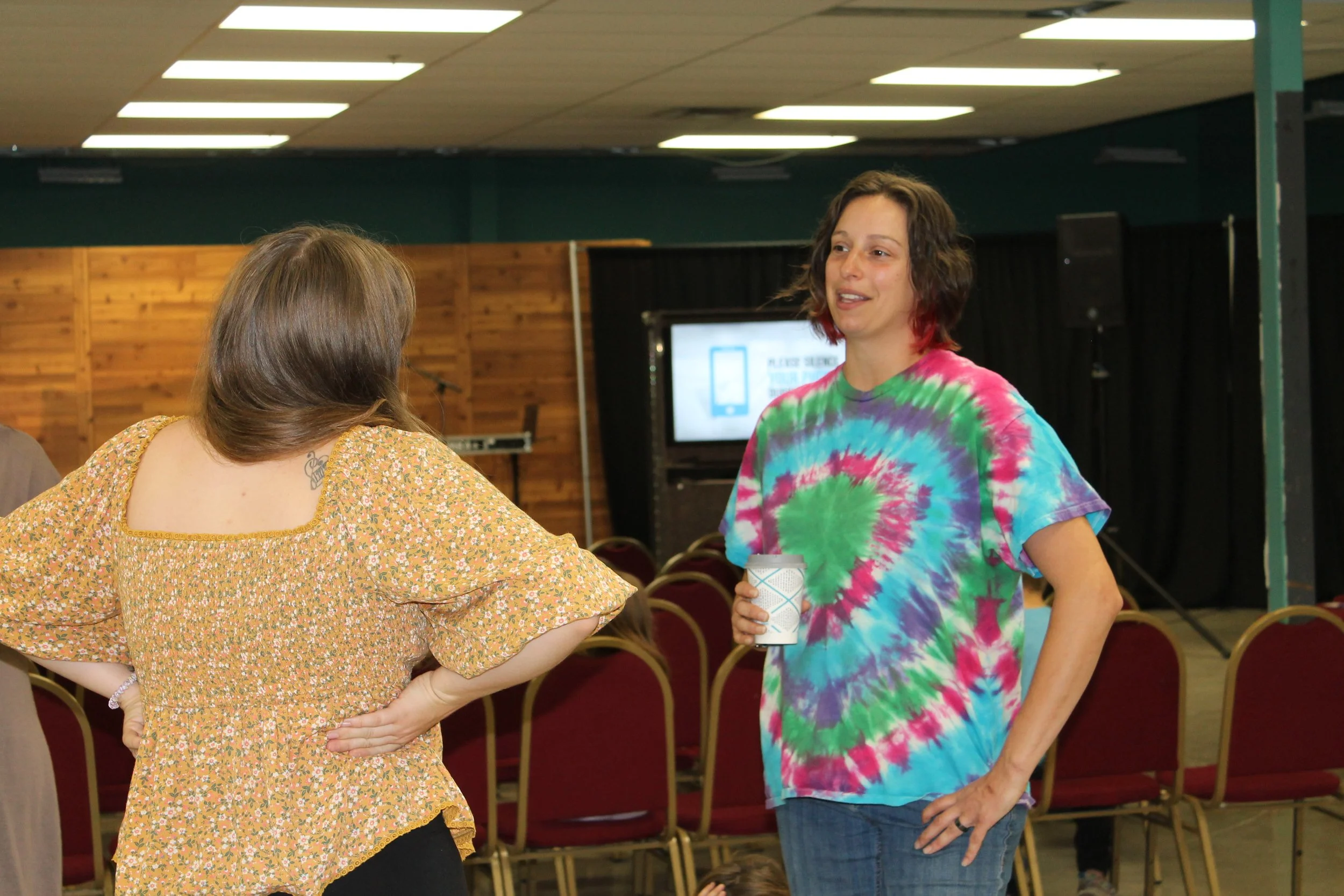 Two women having a conversation in a room with chairs, a wooden wall, and a speaker system. One woman is wearing a yellow floral top with her back to the camera, and the other is wearing a colorful tie-dye shirt, holding a cup, and facing the camera.