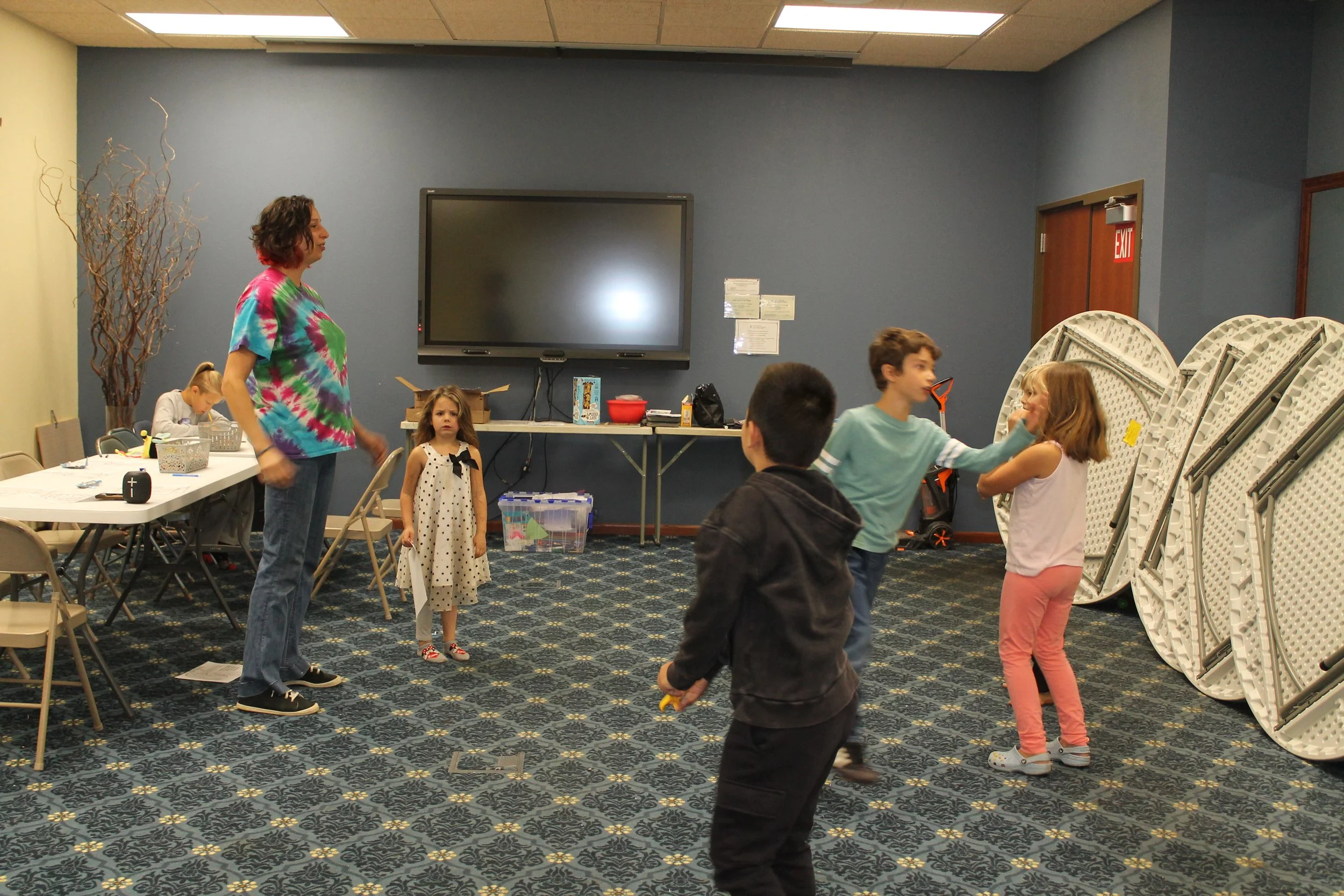 Children playing in a room with blue walls, folding tables, and stacked chairs, while an adult supervises.