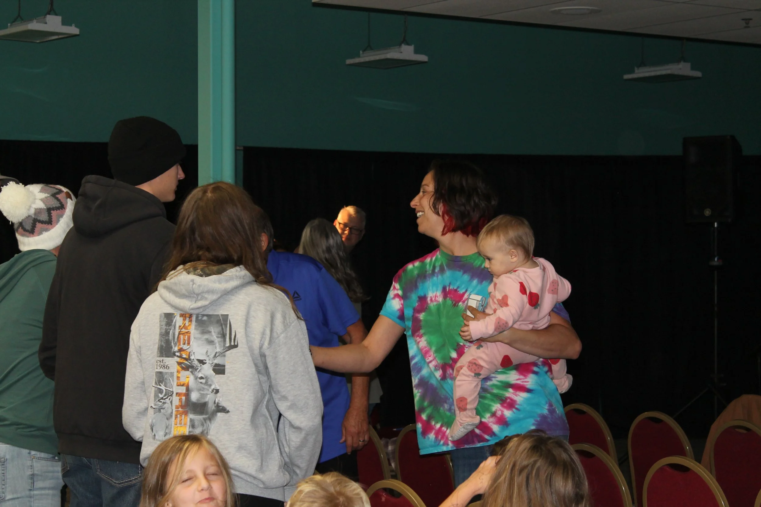 People gathered, engaged in conversation, with a woman holding a young child in a pink outfit. 