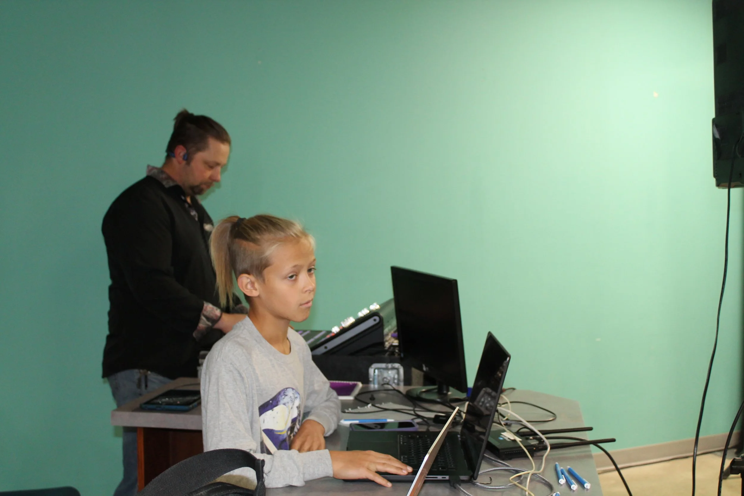 A person working at a desk with multiple computers and equipment while a man stands behind them adjusting something.