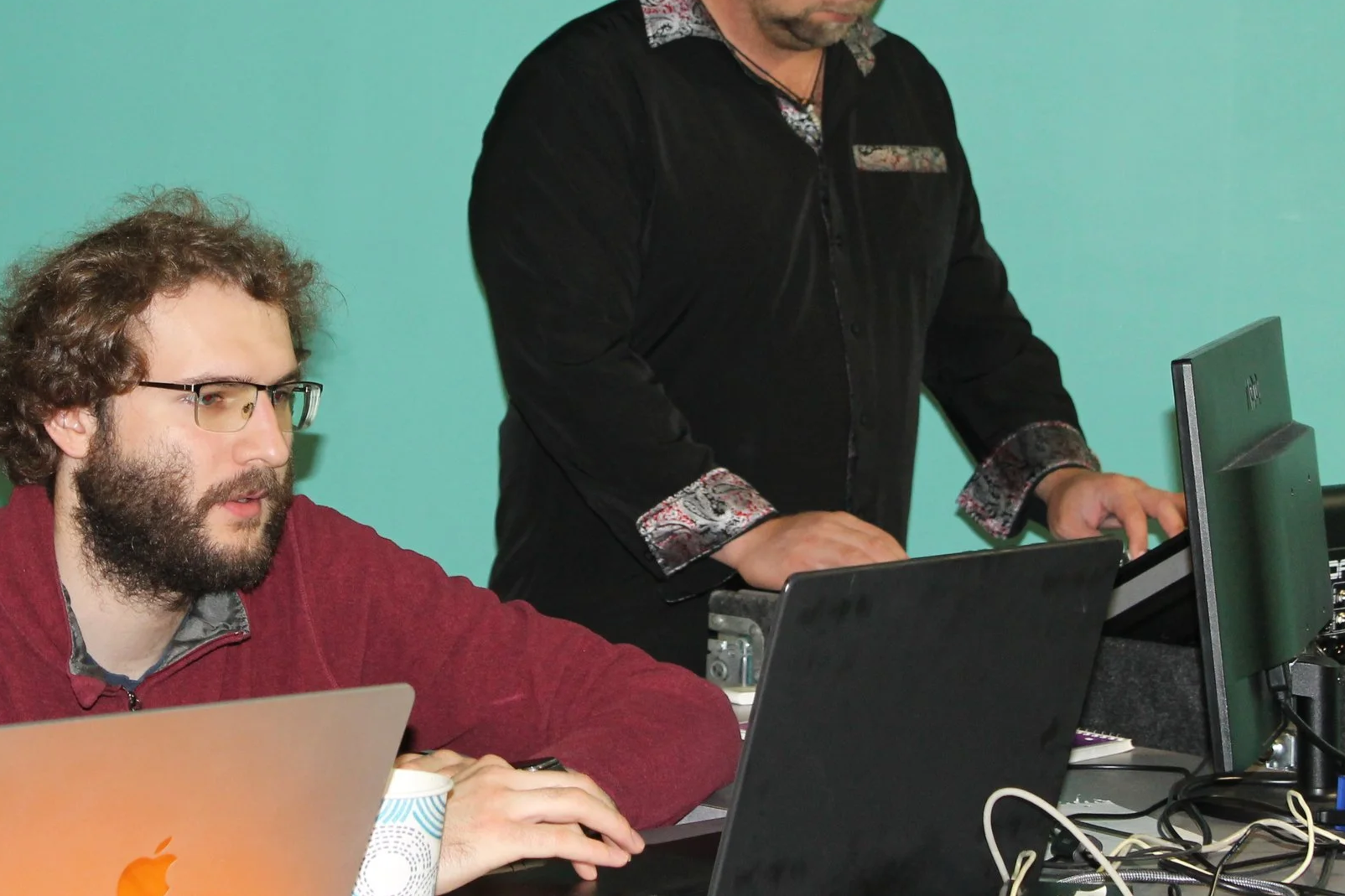 Two men working at a desk with laptops and computer monitors, one wearing glasses and a red sweater, the other standing and operating a keyboard, against a green wall.