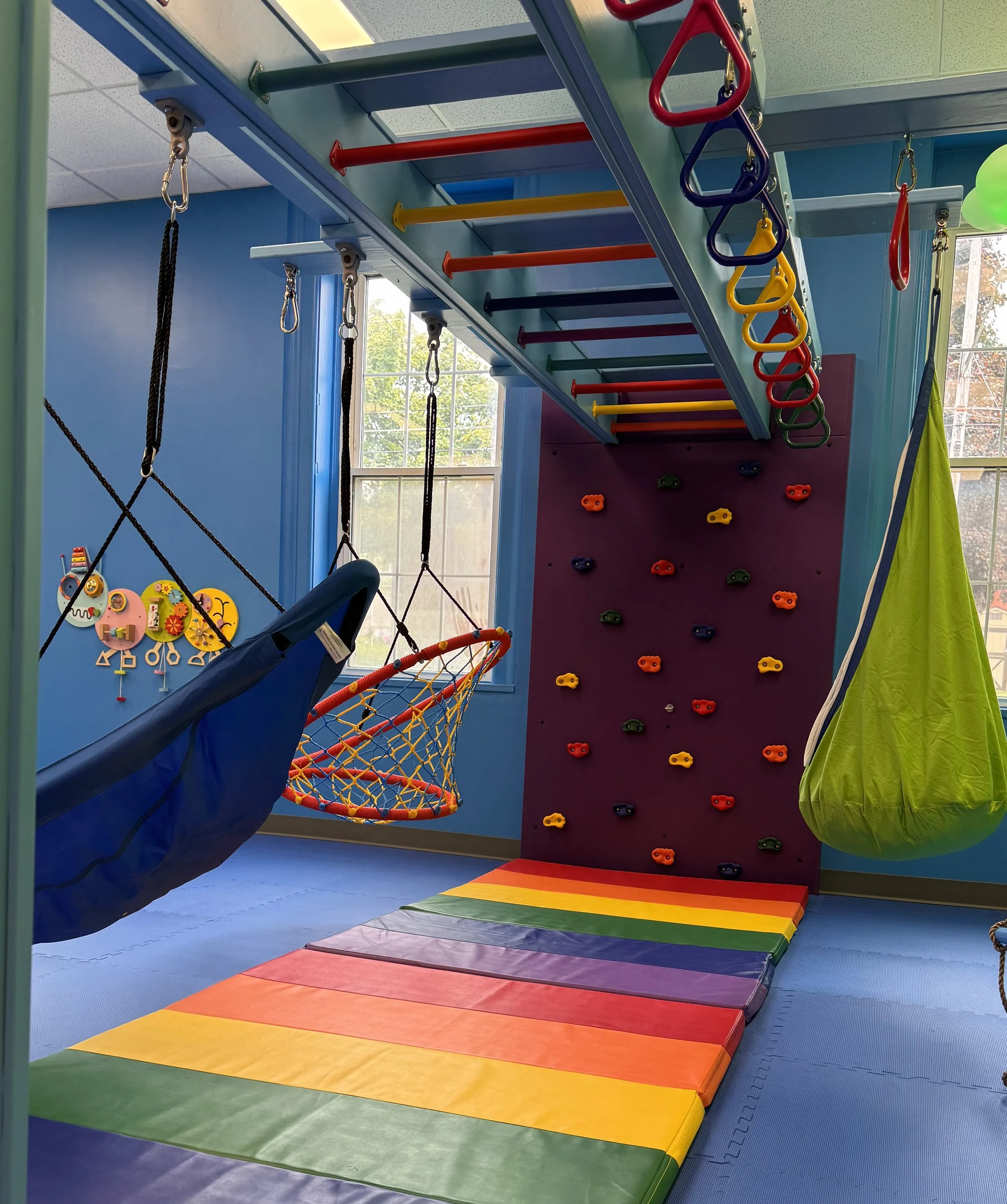 Colorful indoor play area with rainbow-colored mats on the floor, climbing wall with multicolored holds, and various climbing and swinging equipment for children.
