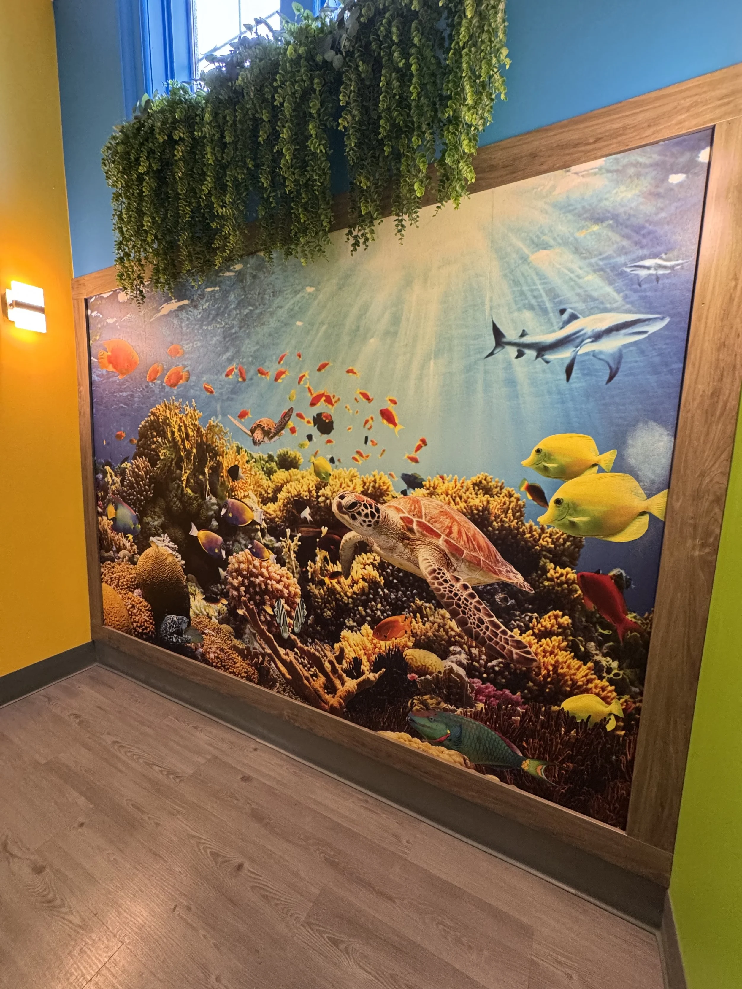 A colorful underwater scene mural with various fish, a sea turtle, coral reefs, and sunlight filtering through water, framed with a wooden border, mounted on a wall with yellow and green sections and a wooden floor.
