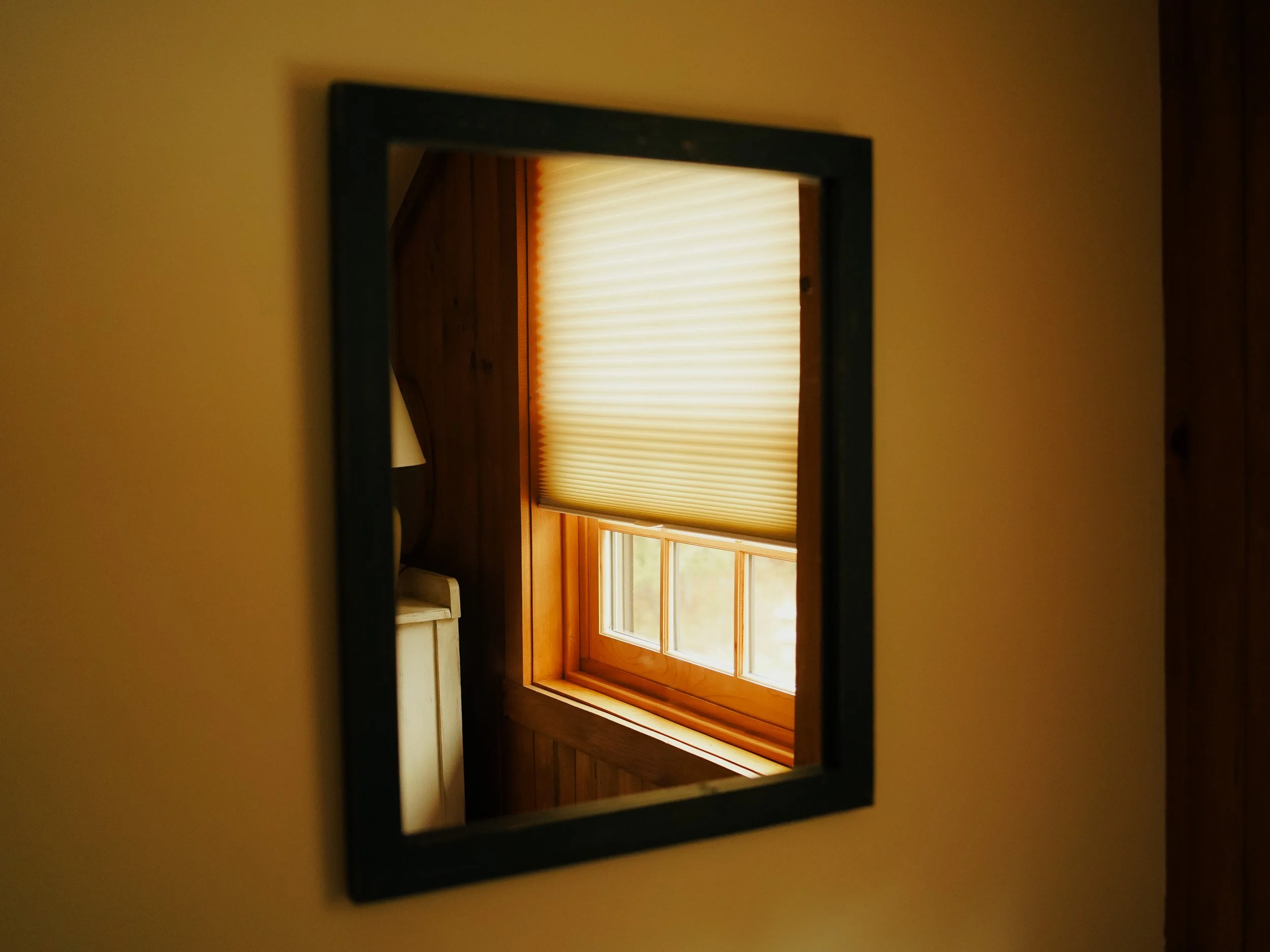 Reflection in a small rectangular mirror shows a window with a beige window shade, wooden wall paneling, and a white cabinet.