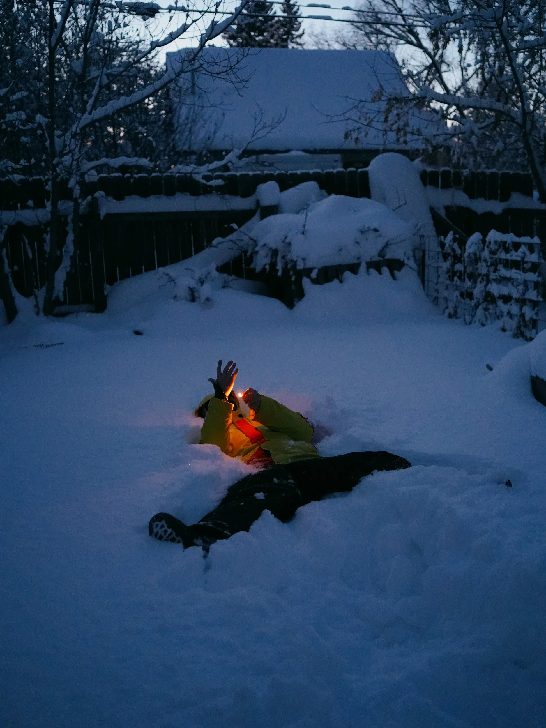 Person lying on snow in backyard, wearing a yellow jacket and black pants, holding a lit lighter, with snow-covered trees and house in background during dusk.