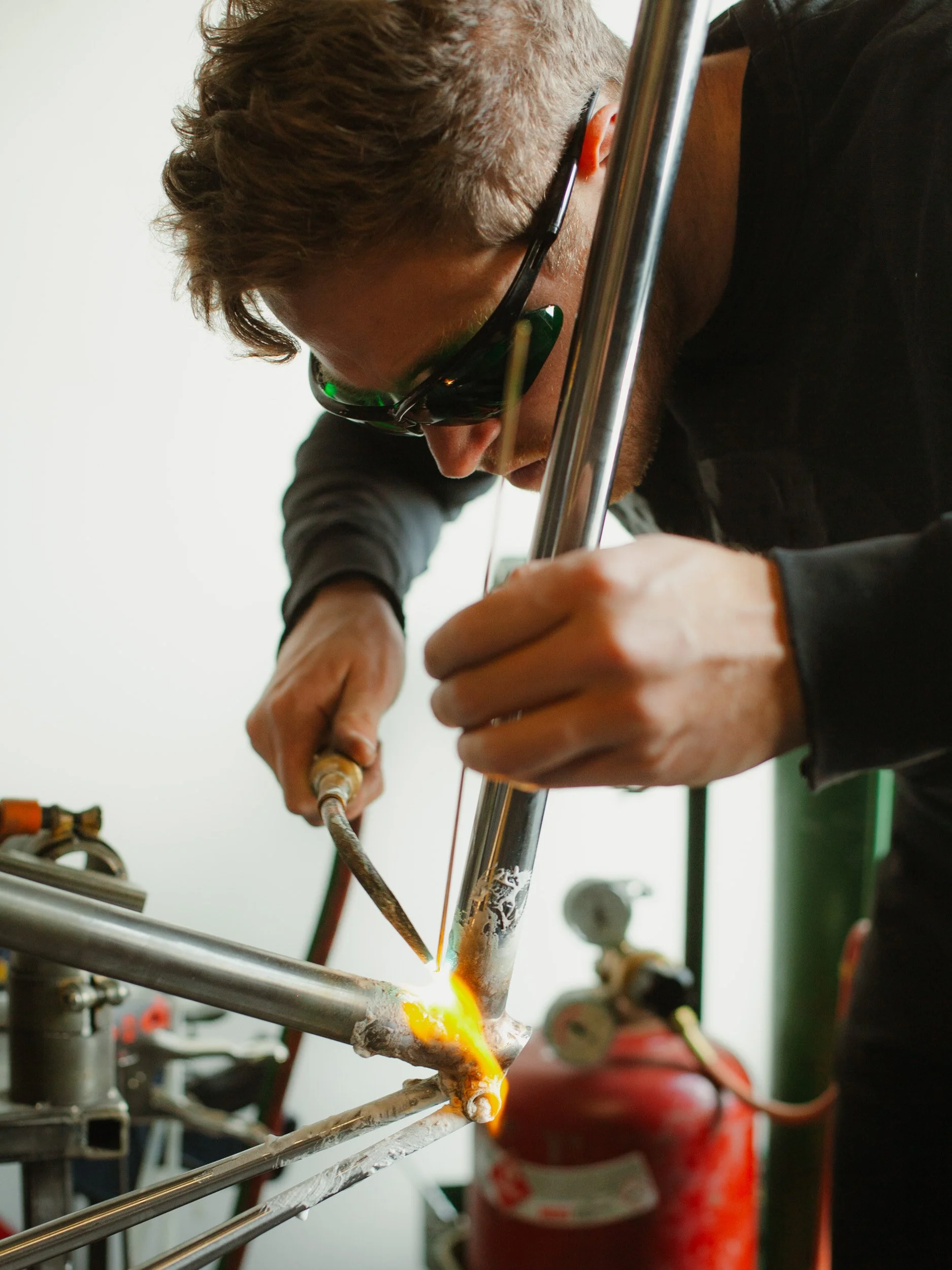 A man welding a metal bicycle frame in a workshop, wearing safety glasses and using a welding torch.