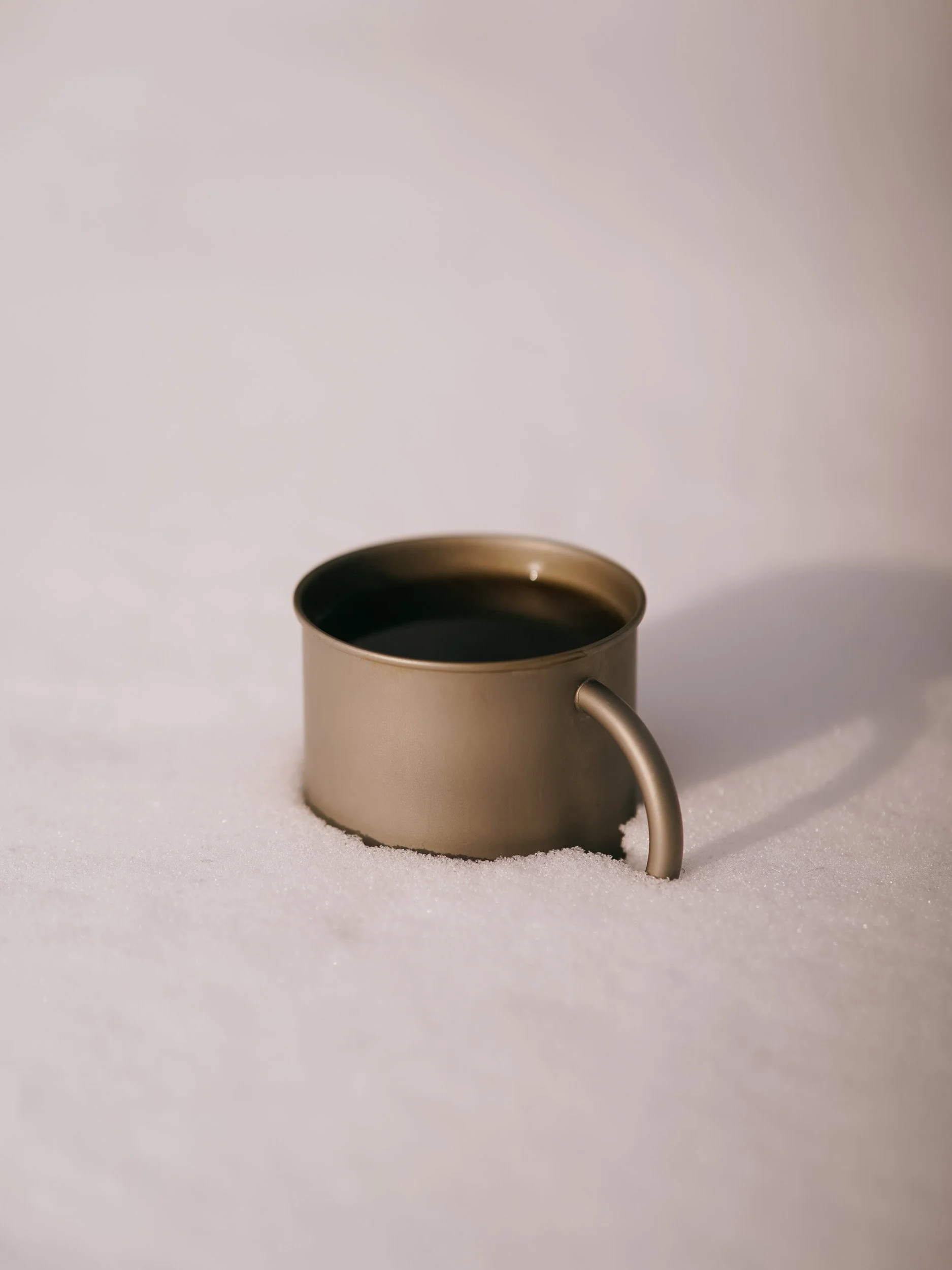 A metal cup filled with dark liquid, placed on snow with a soft shadow cast to the right.