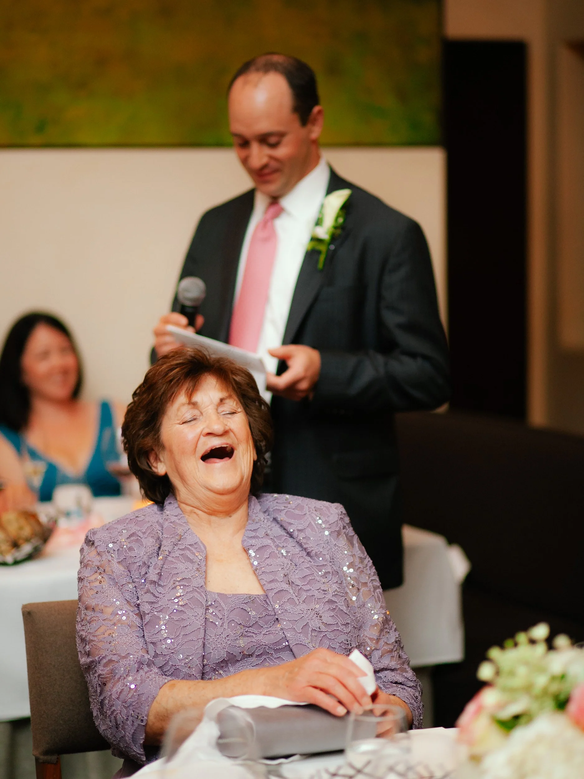 A woman in a lavender lace dress is laughing and appears to be crying tears of joy, while a man in a suit and pink tie stands behind her, holding a microphone and reading from a paper during a celebration event, with other women smiling in the backgr