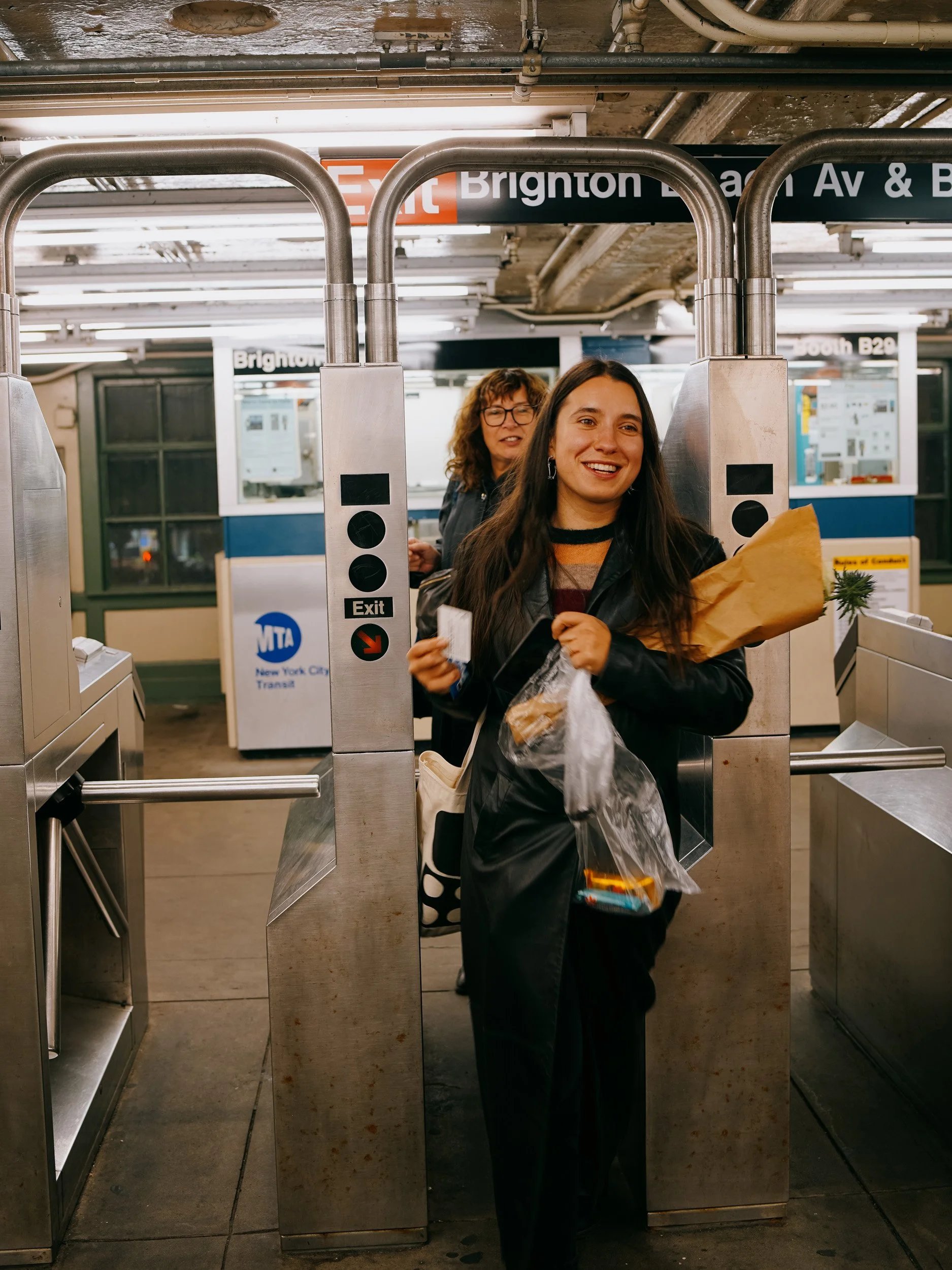 Two women smiling at a subway entrance, holding groceries, with the New York City subway station sign in the background.