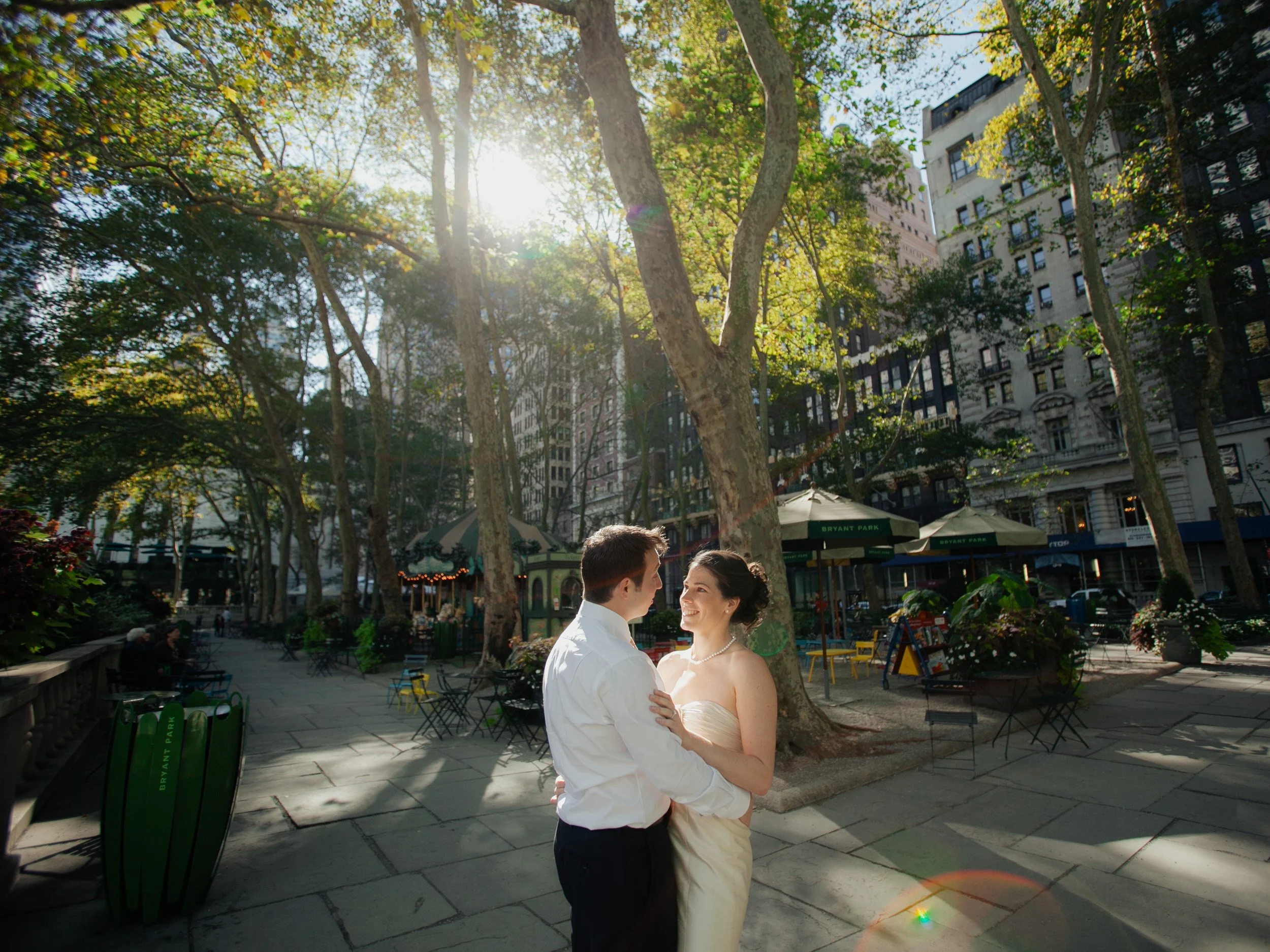 A couple dancing in Bryant Park on a sunny day with trees, buildings, and park benches in the background.