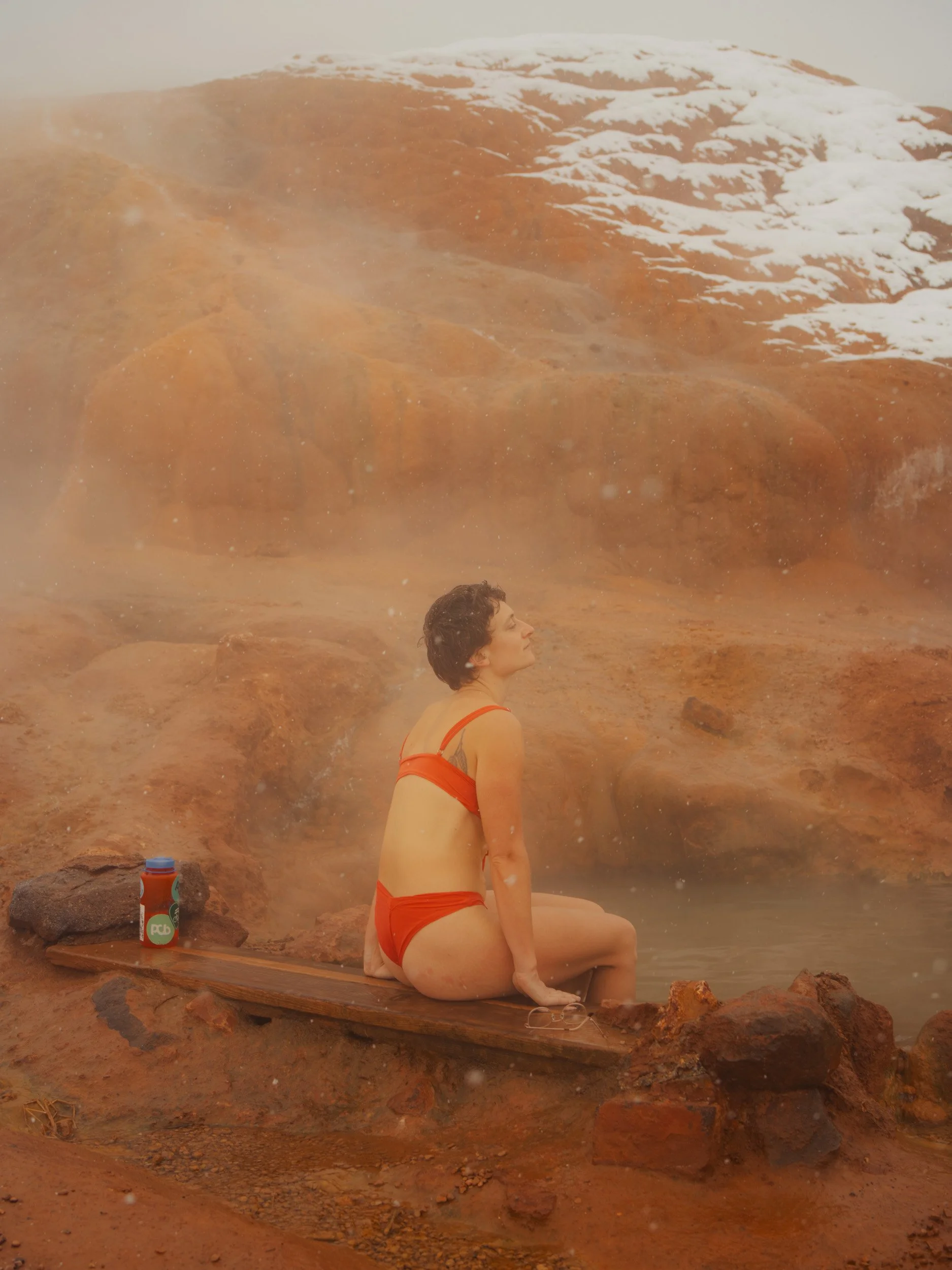 A woman in a red bikini sitting on a wooden platform in a hot spring, with steam rising around her and snow on the rocky hillside in the background.