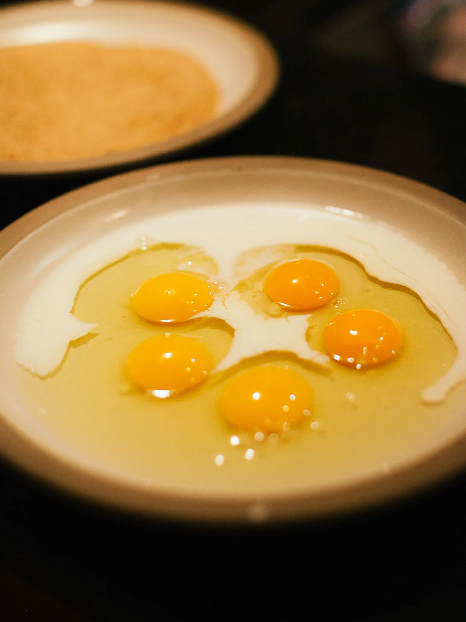 Five raw egg yolks in a white bowl, with a plate of seasoned bread crumbs in the background.