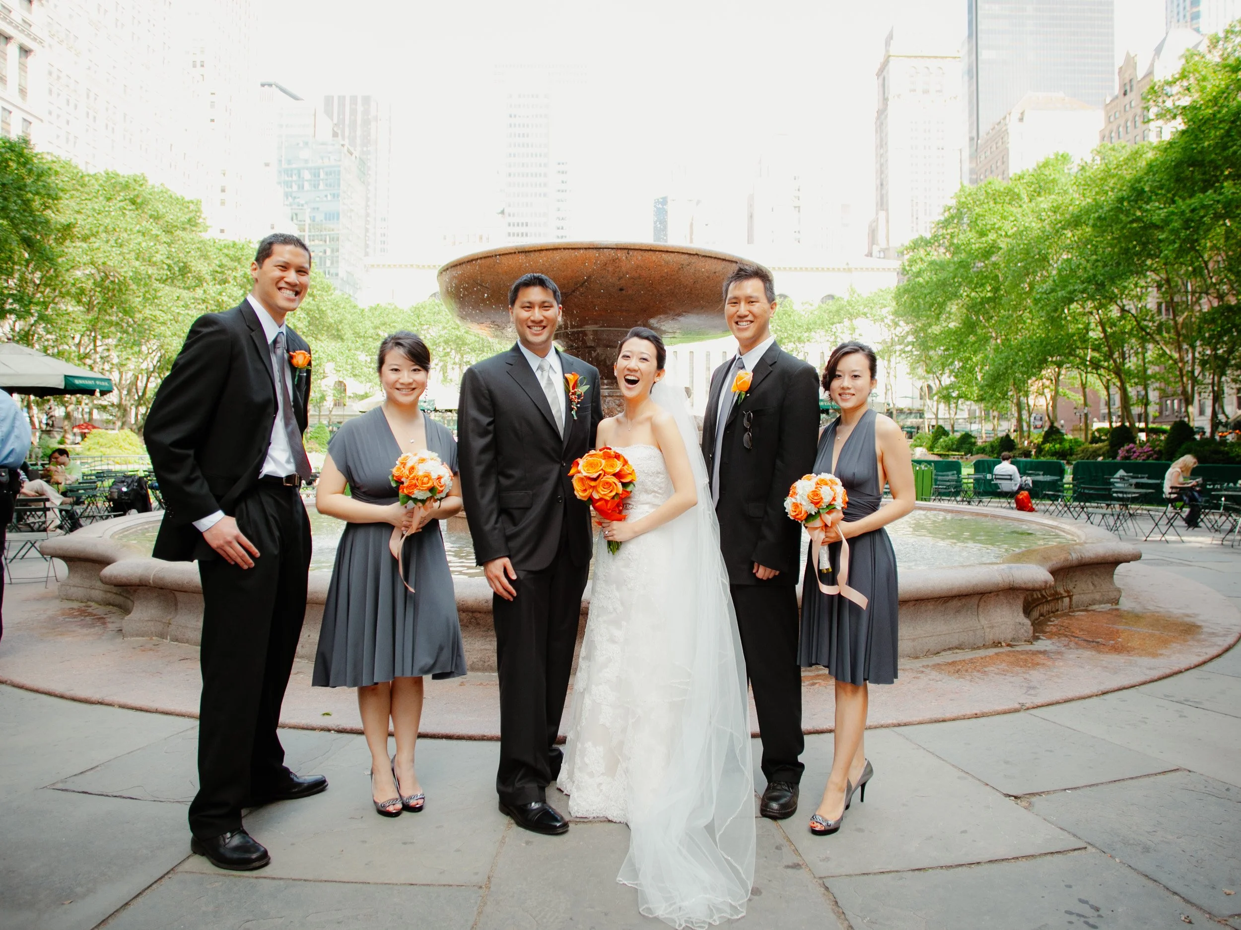 A group of seven people, including a bride and groom, standing in front of a fountain in an urban park, smiling, dressed in wedding attire with the bride in a white gown and the groom in a dark suit, surrounded by bridesmaids and groomsmen holding bo