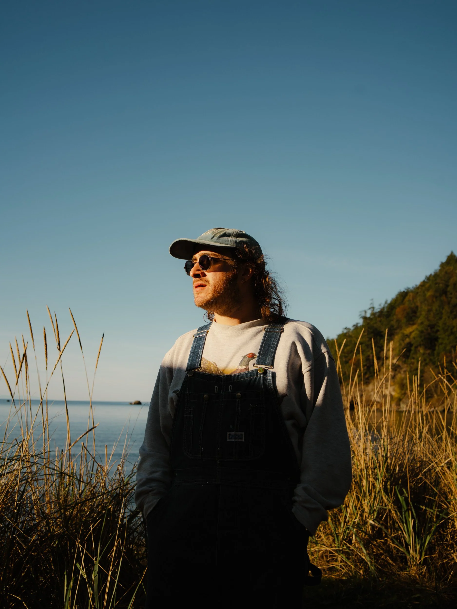A person with long hair wearing sunglasses, a gray cap, a gray sweatshirt, and overalls stands outdoors near a body of water, with tall grass and a hill in the background.