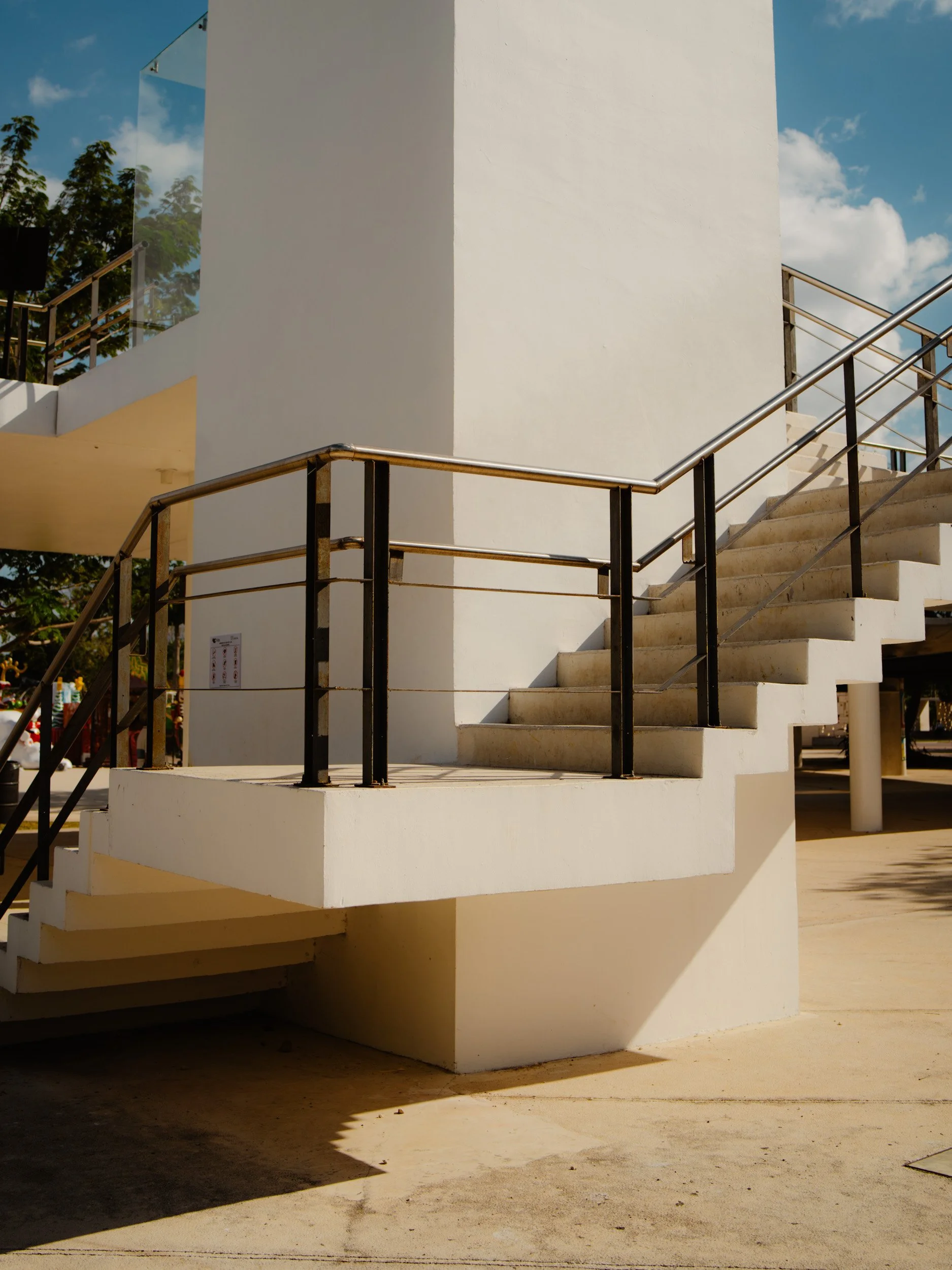 Exterior view of a white staircase with metal handrails, leading up around a white building under a partly cloudy sky.