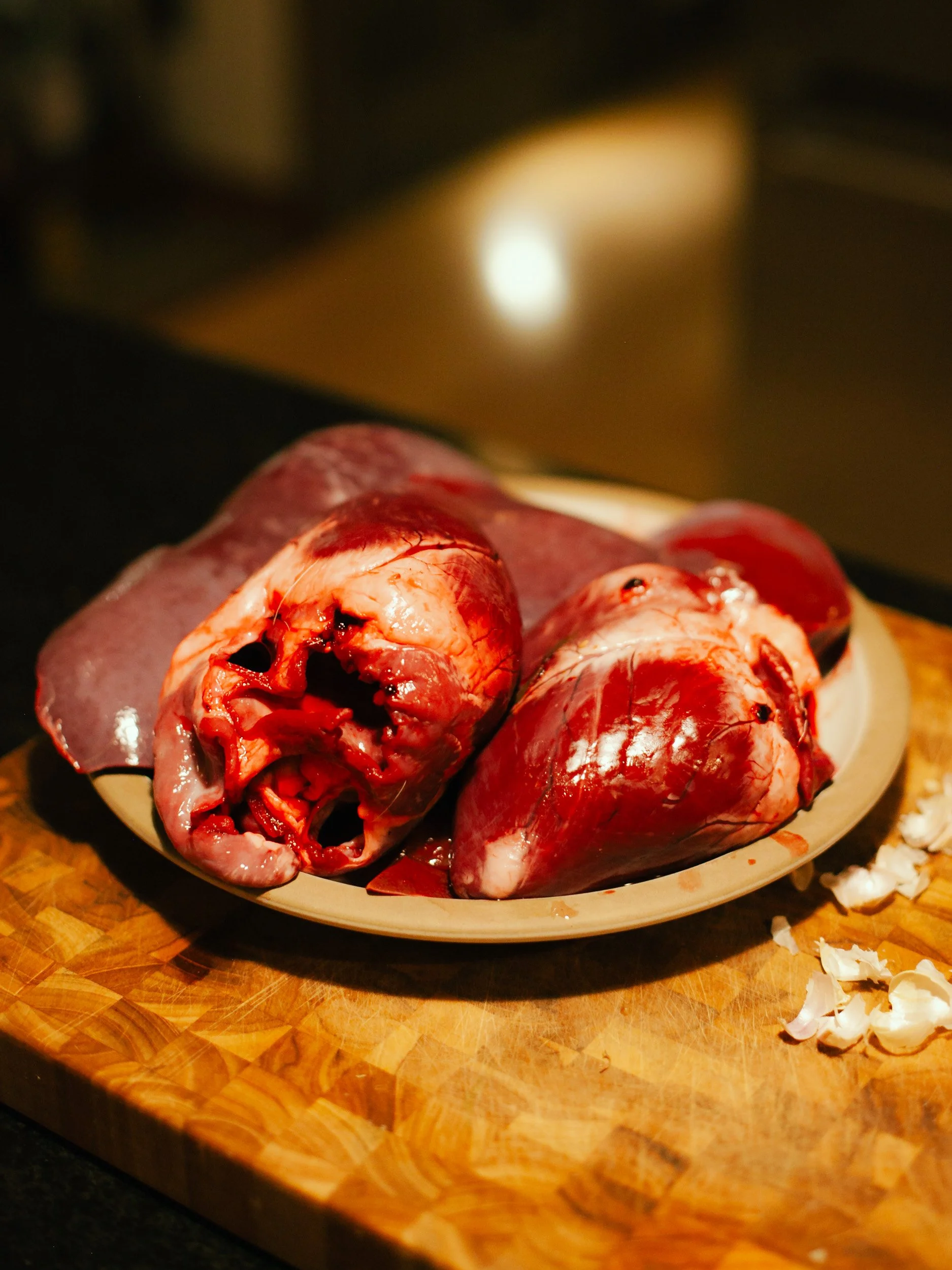 Raw animal hearts on a plate with garlic cloves on a wooden cutting board.