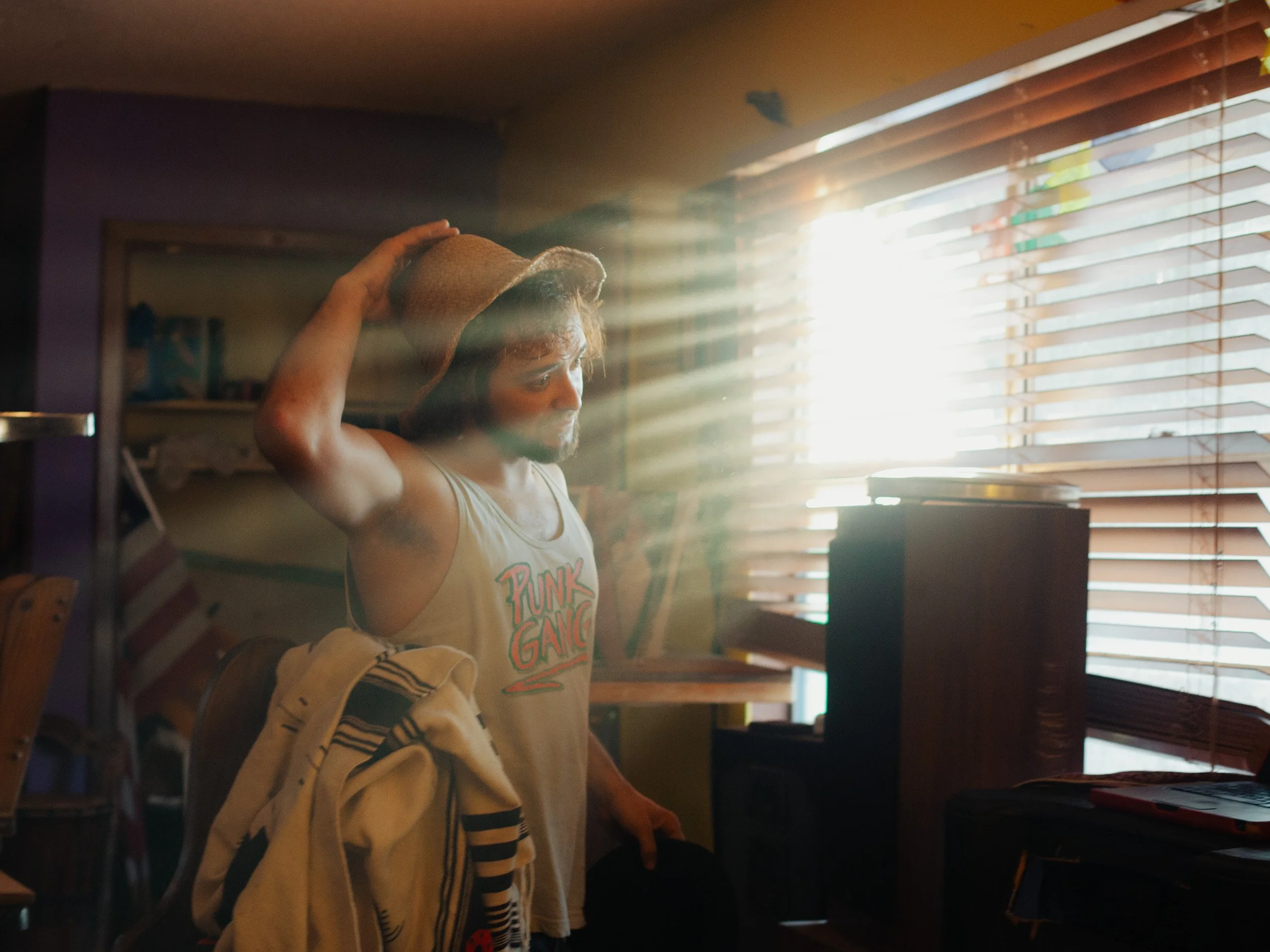 A man with curly hair and a beard wearing a sleeveless shirt with 'Punk Gang' text, a straw hat, and holding another hat, standing indoors with sunlight streaming through window blinds.
