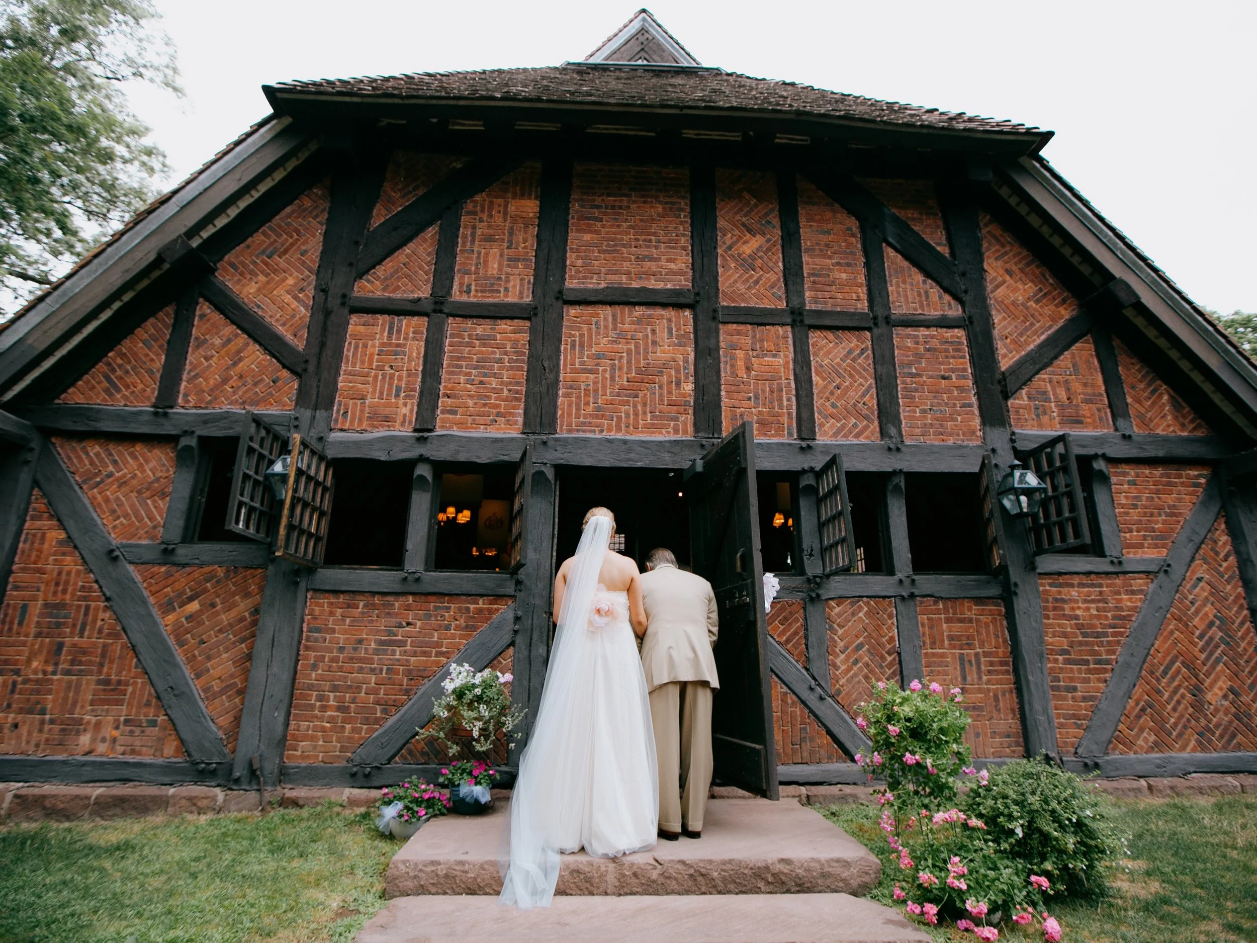 A bride in a white wedding dress and veil walking into a rustic brick building with a man in a beige suit, surrounded by pink and purple flowers.