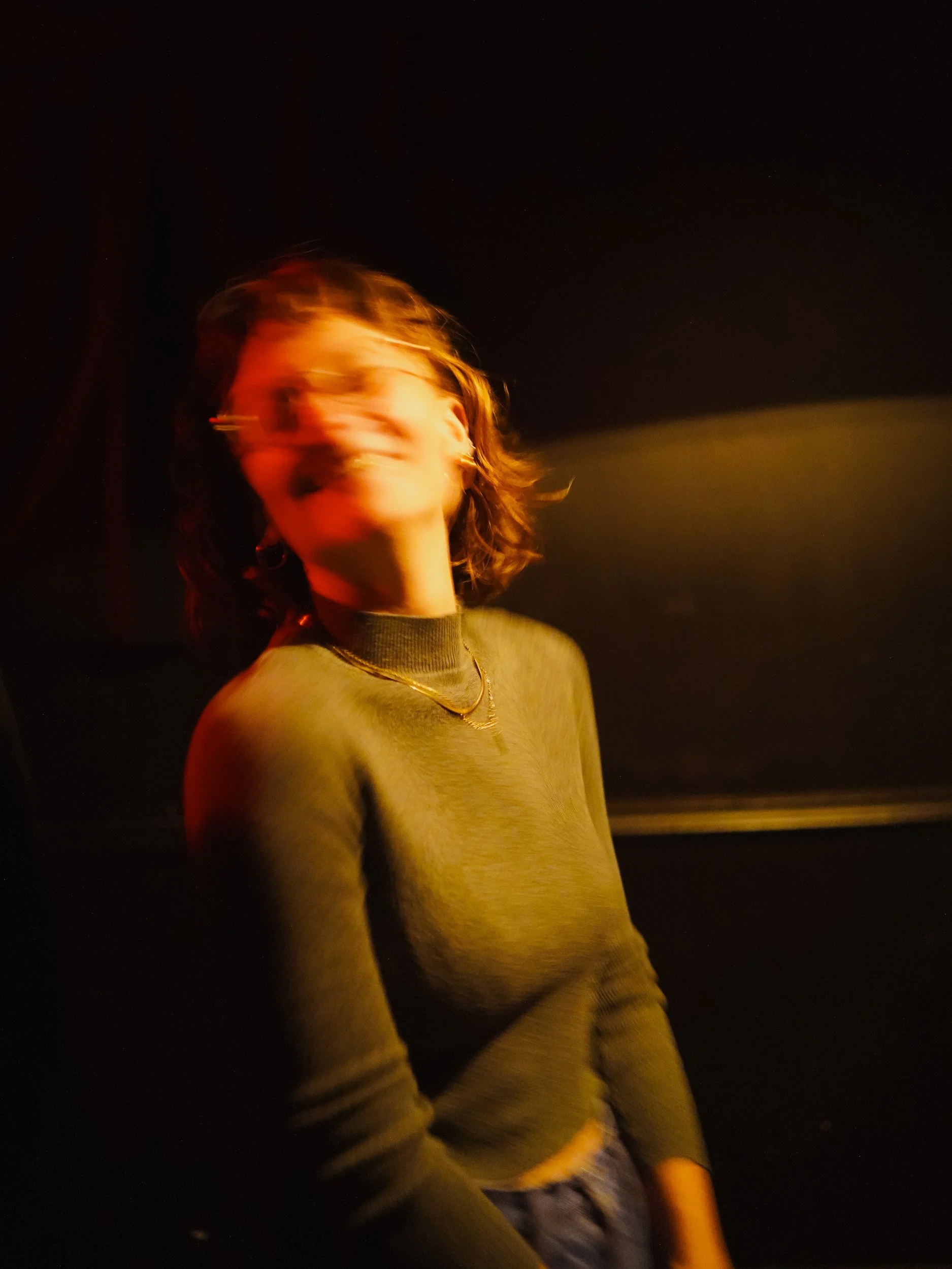 Blurred image of a woman with shoulder-length hair, laughing or smiling, wearing a green sweater and layered necklaces, in a dark setting with warm lighting.