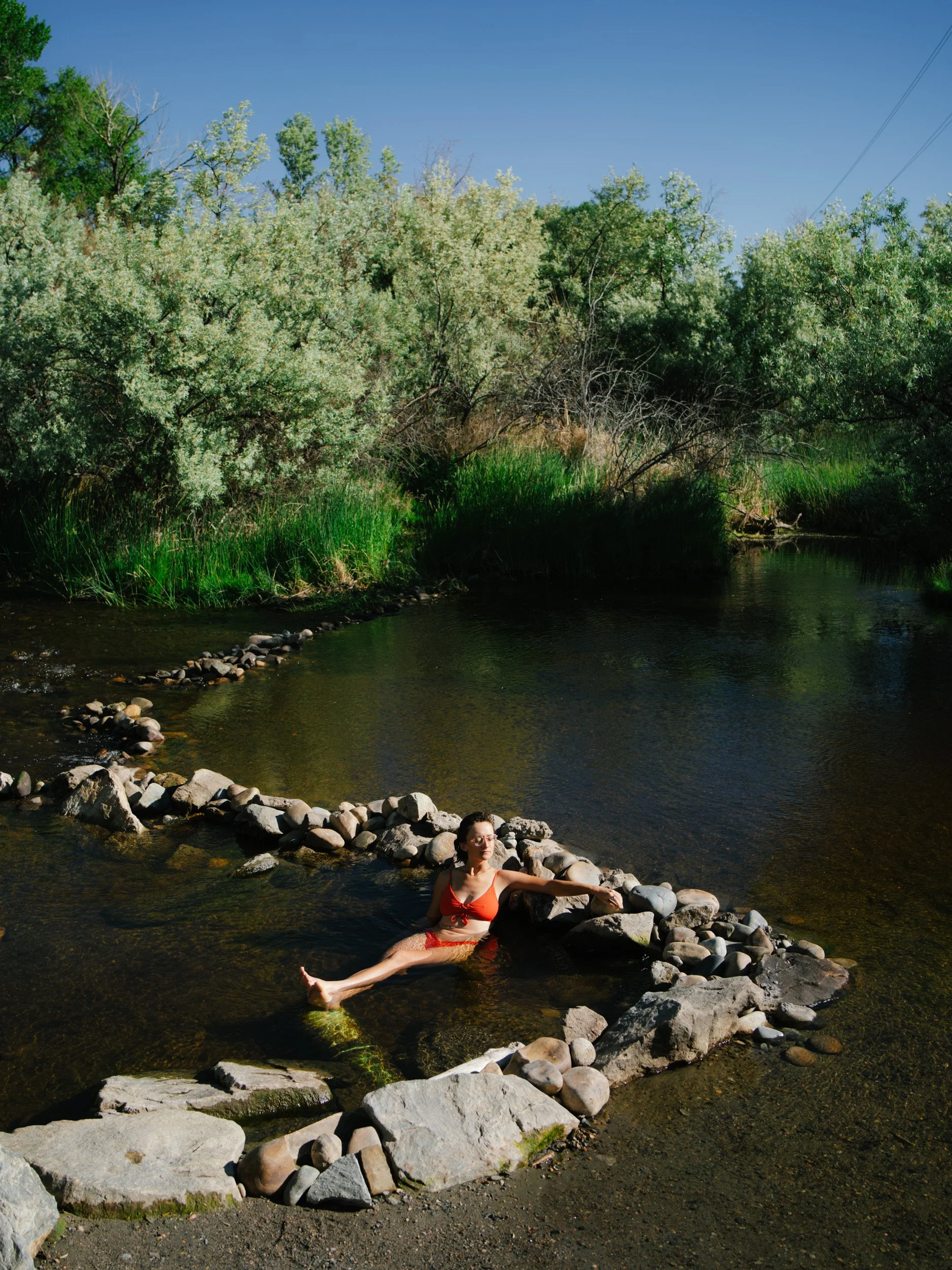 A woman in a red swimsuit relaxing in a rocky section of a river surrounded by green trees and bushes under a clear blue sky.