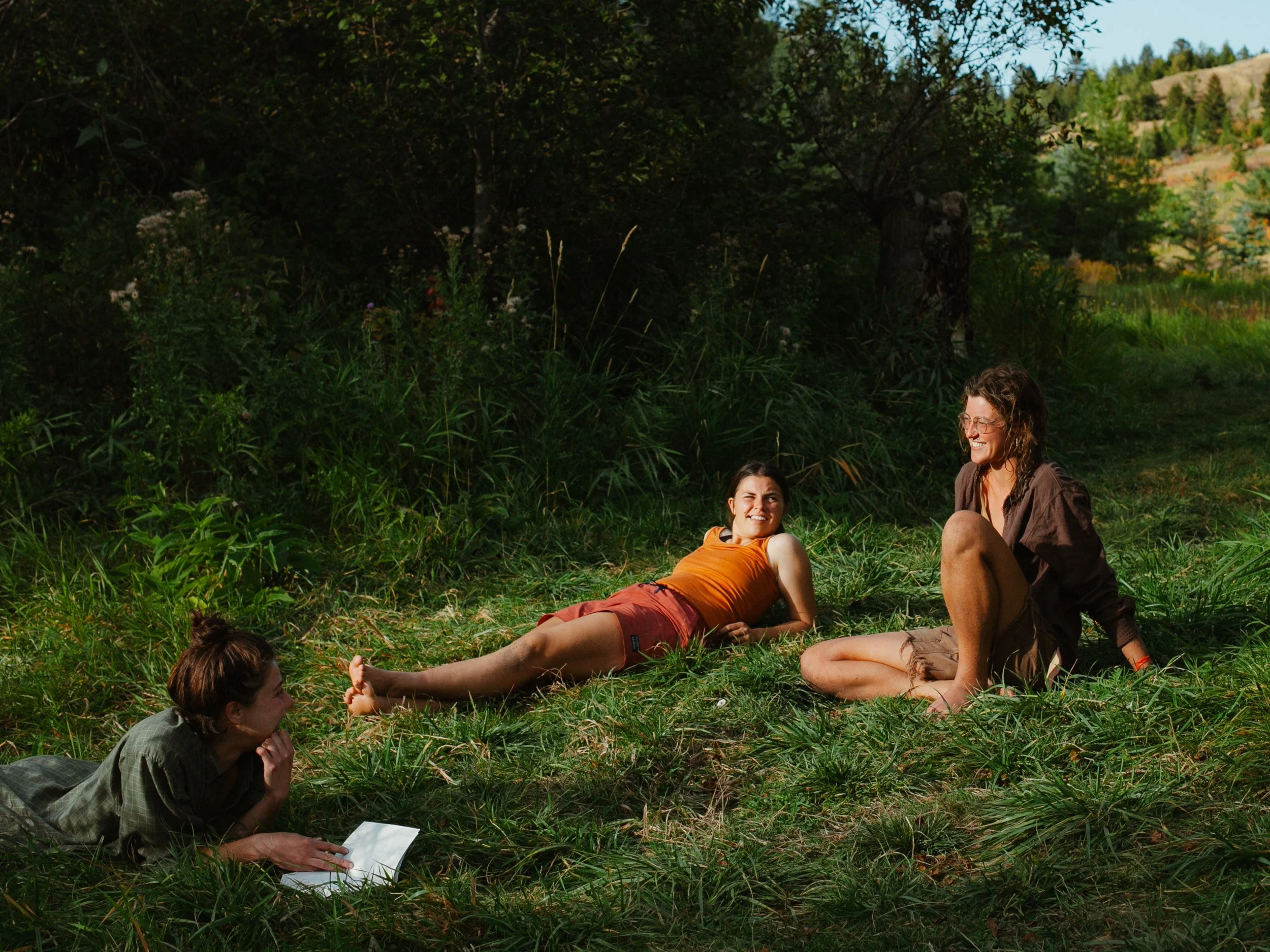 Three women are relaxing outdoors in a grassy area with trees and hills in the background. One woman is lying on her back, another is sitting with her knee bent, and the third is lying face down with a notebook nearby.