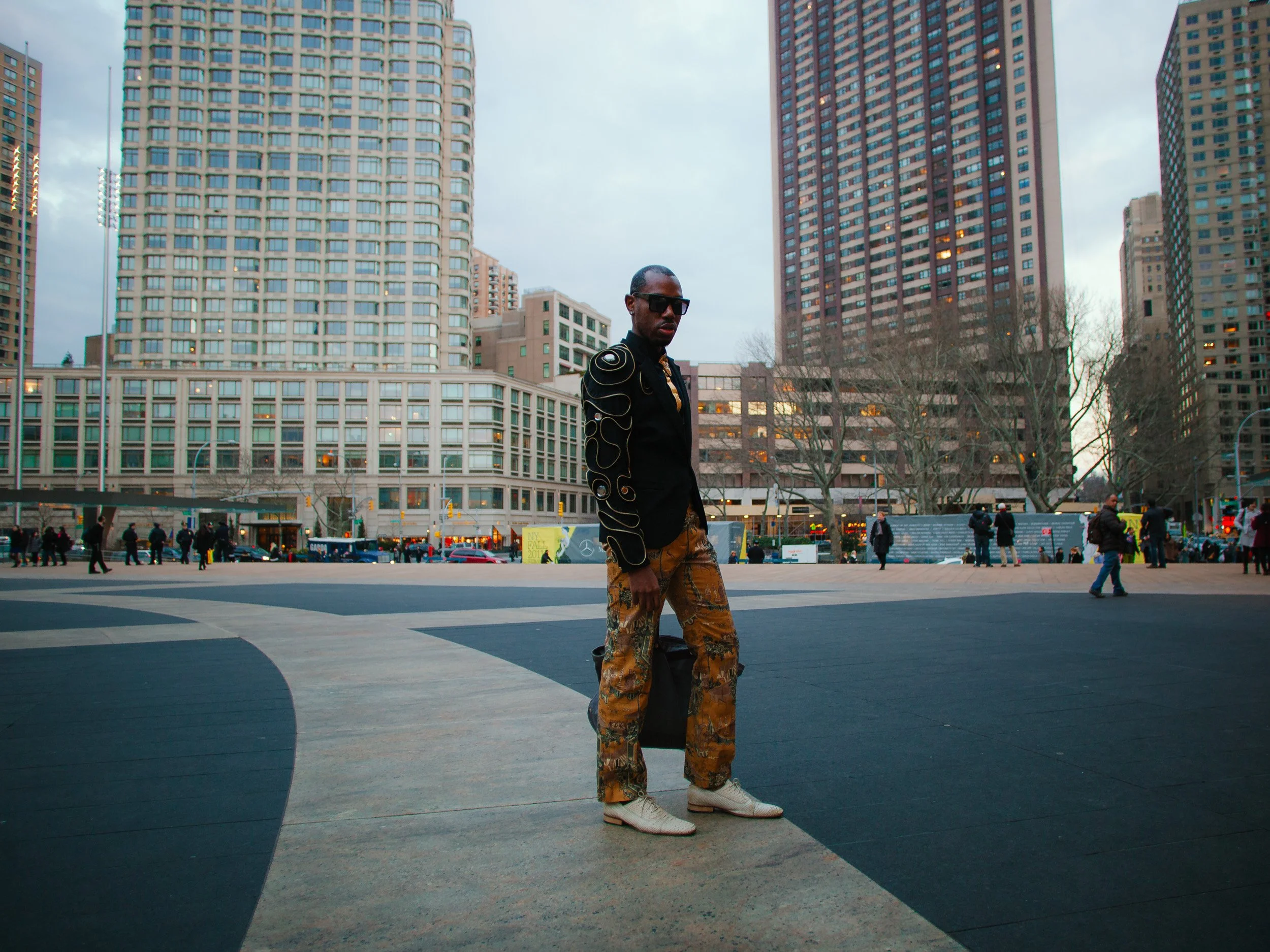 A man with sunglasses wearing a black jacket with gold designs, camouflage pants, and white shoes standing in an urban park with tall buildings and trees in the background.