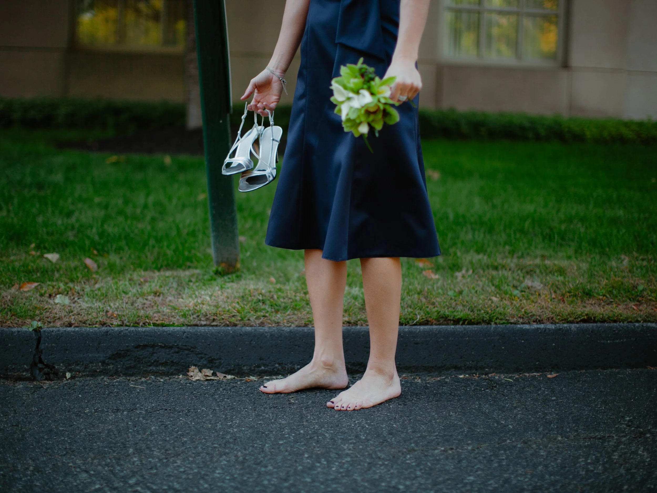 Person standing barefoot on the sidewalk holding a bouquet of flowers in one hand and a pair of silver high heels in the other, with a grassy area and building in the background.