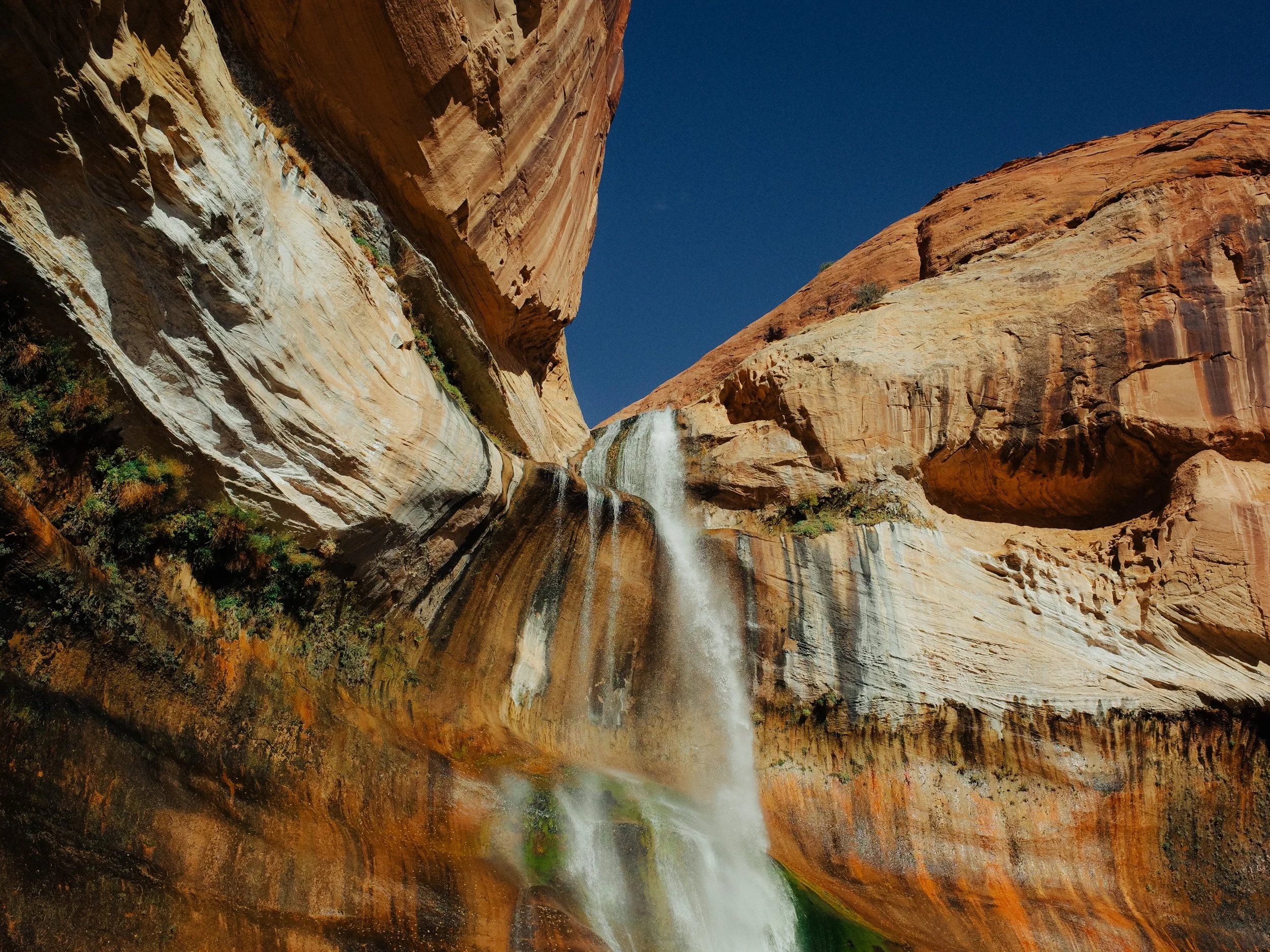A waterfall flowing down a narrow canyon with reddish rock walls and a clear blue sky above.
