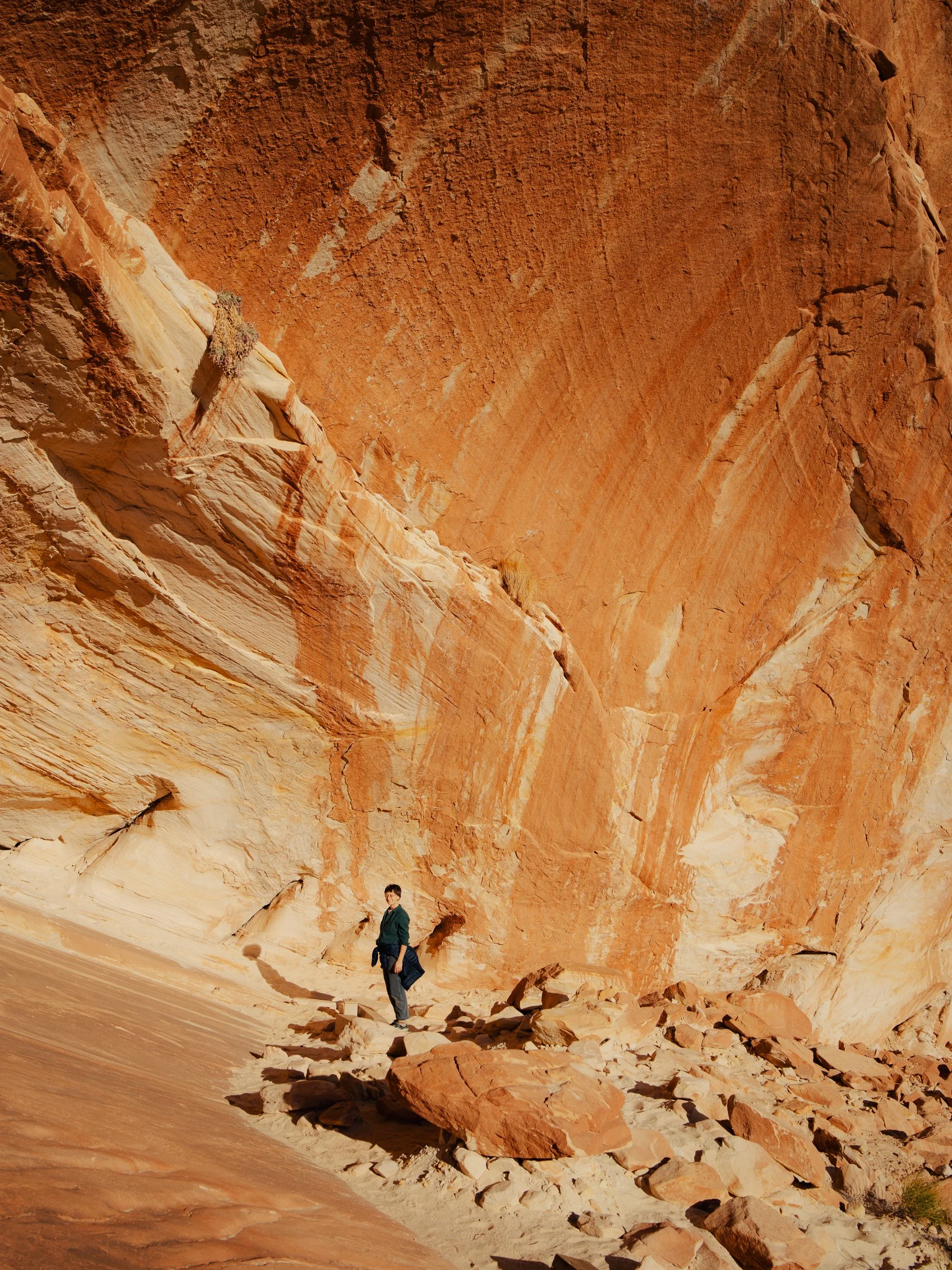 A person standing on rocky desert terrain at the base of a large red sandstone cliff.