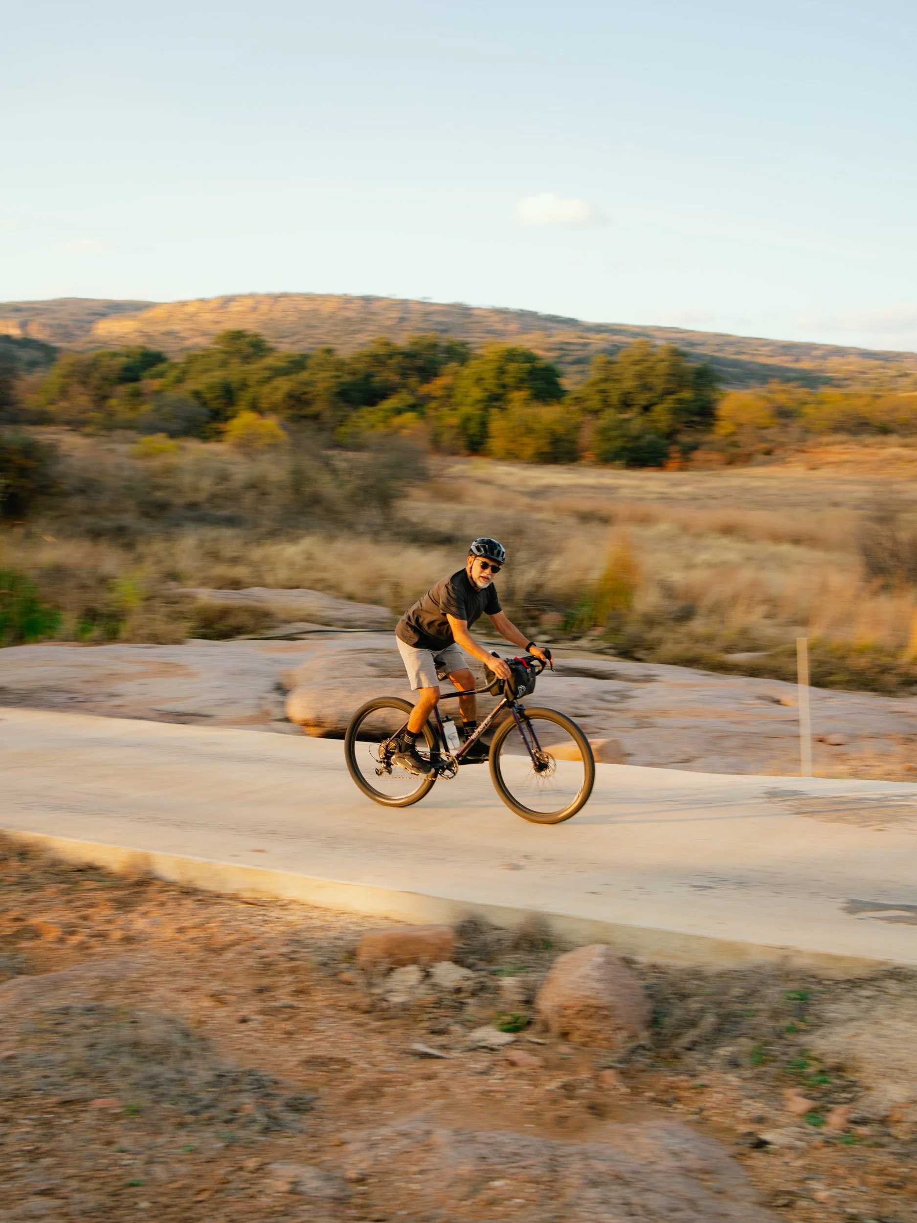 A man riding a mountain bike on a dirt trail in a hilly, open landscape with trees and dry grass, during sunset.