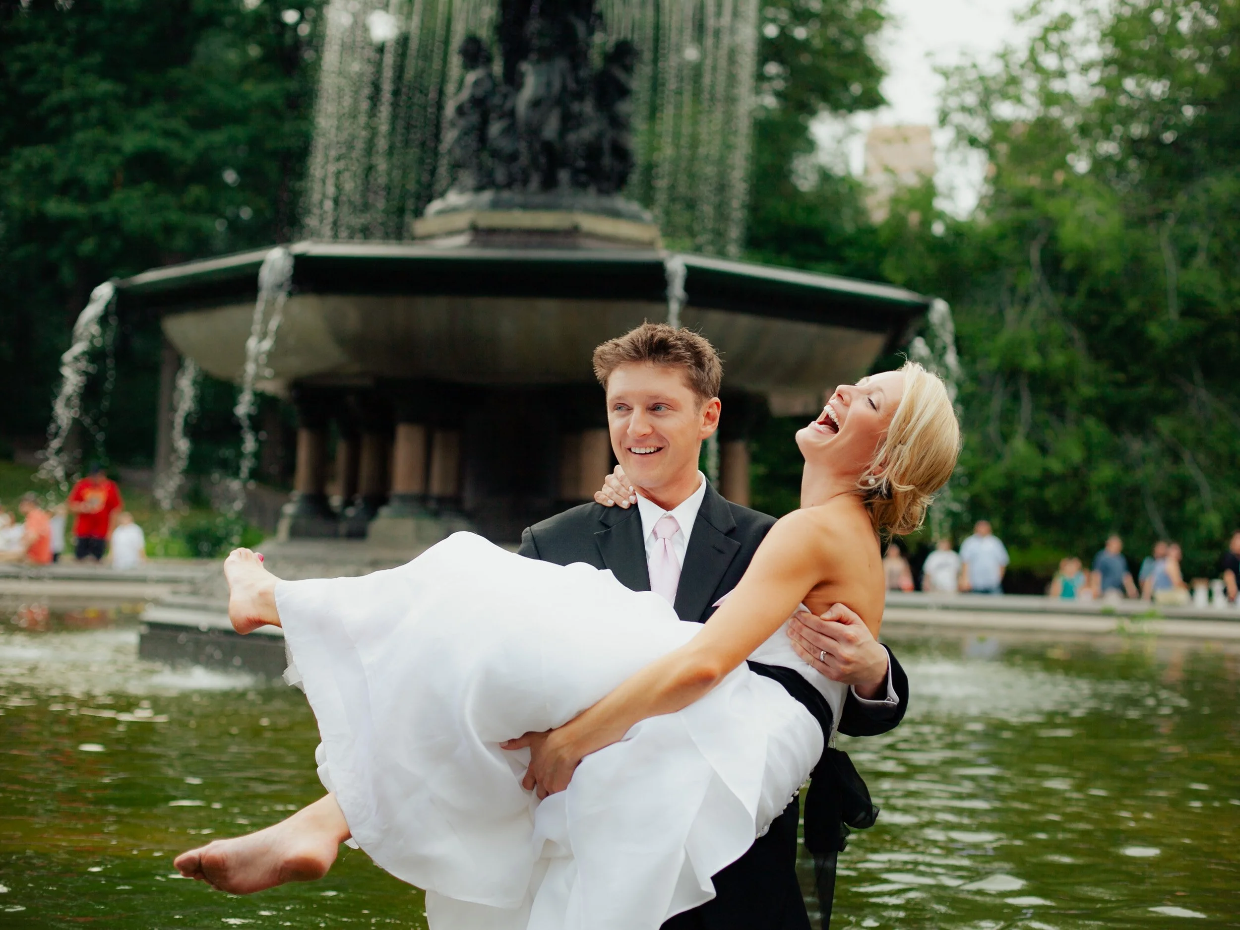 A joyful couple in wedding attire, the man in a black suit carrying the woman in a white dress, next to a fountain in a park.
