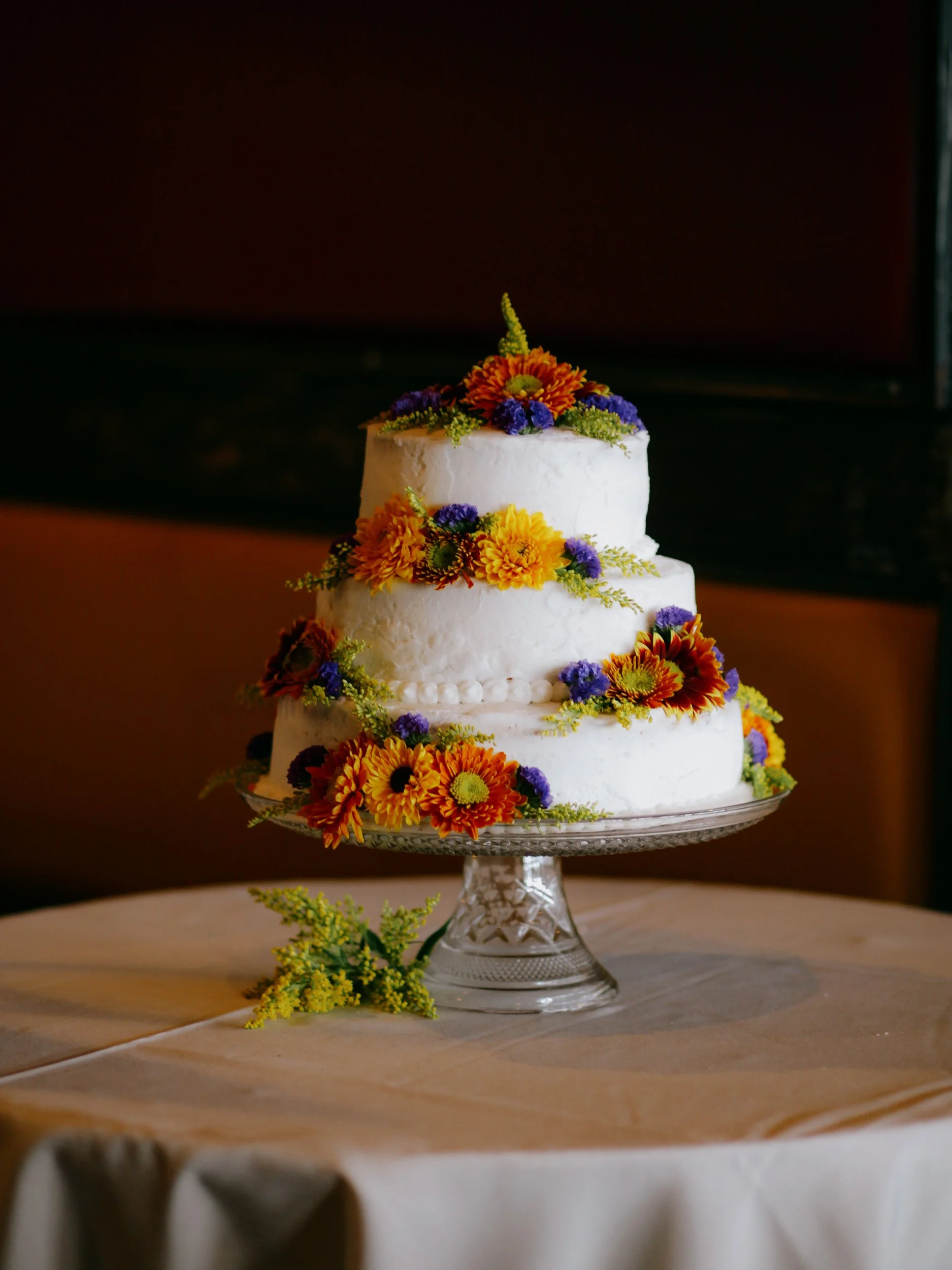 Three-tiered white wedding cake decorated with orange, yellow, and purple flowers on a glass cake stand, sitting on a table with a beige tablecloth.