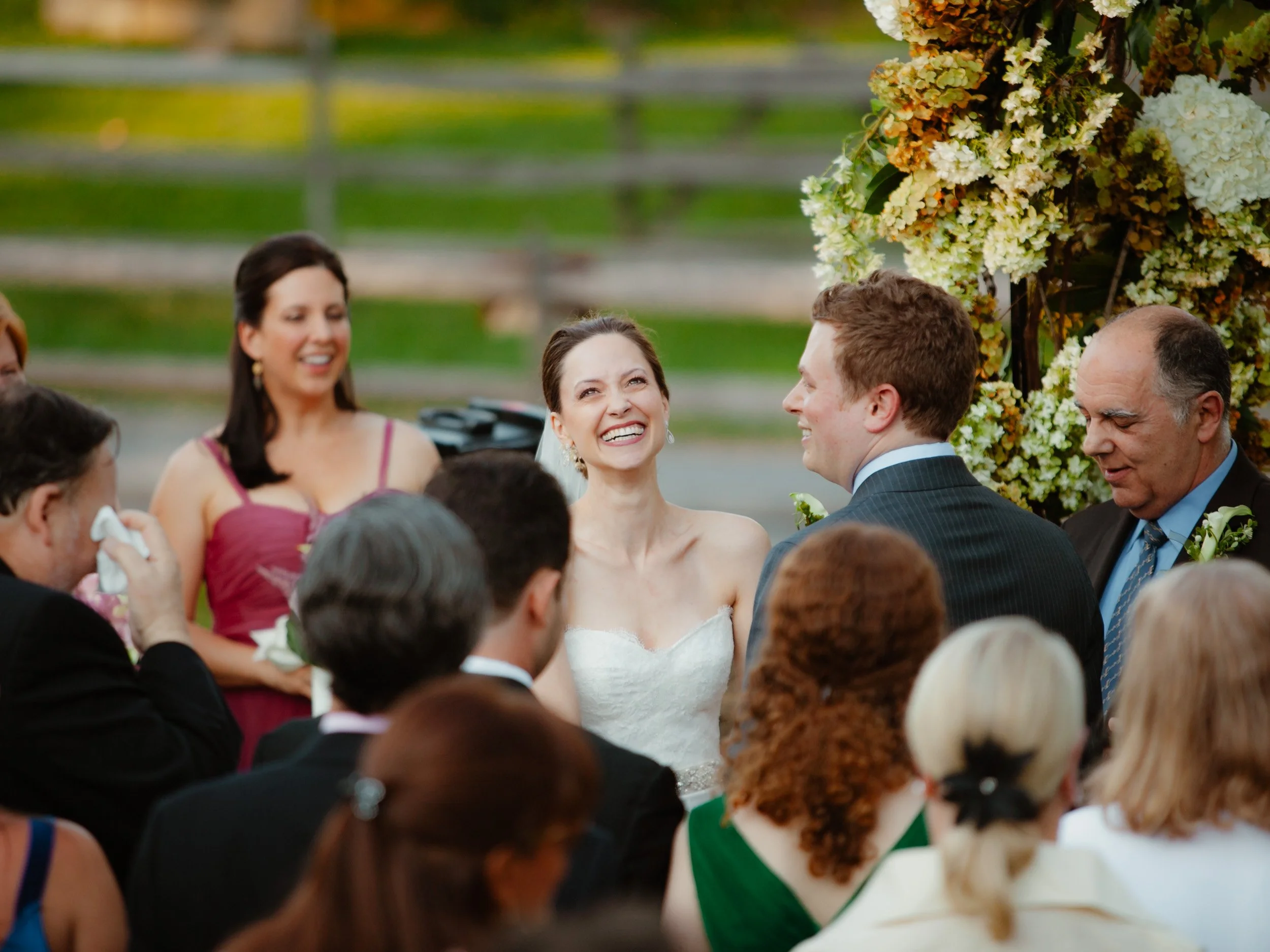 A bride and groom exchanging vows at a wedding ceremony, surrounded by friends and family, outdoors with a floral backdrop.