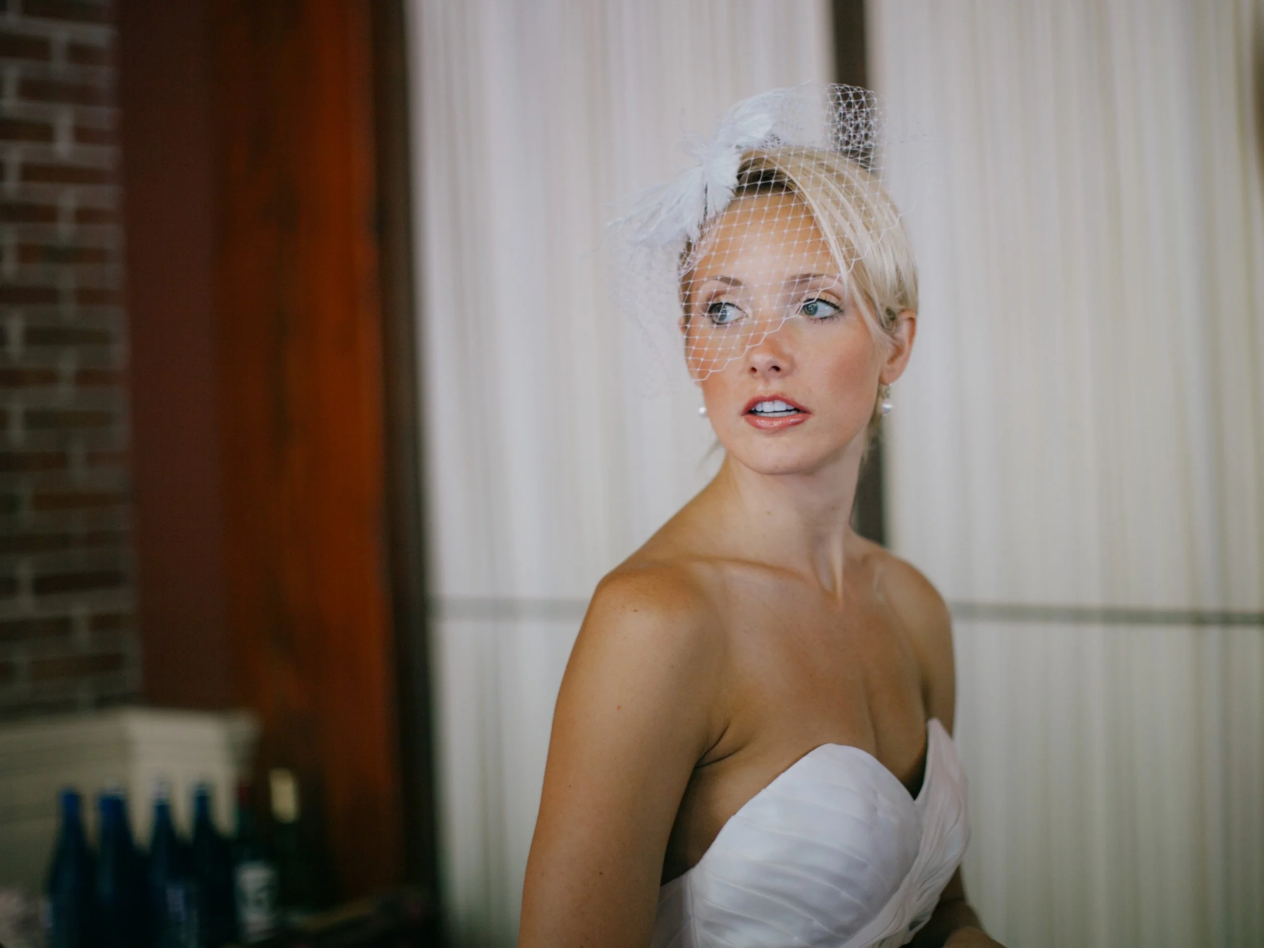 A blonde woman in a wedding dress with a birdcage veil looking to her right indoors with a focused expression.