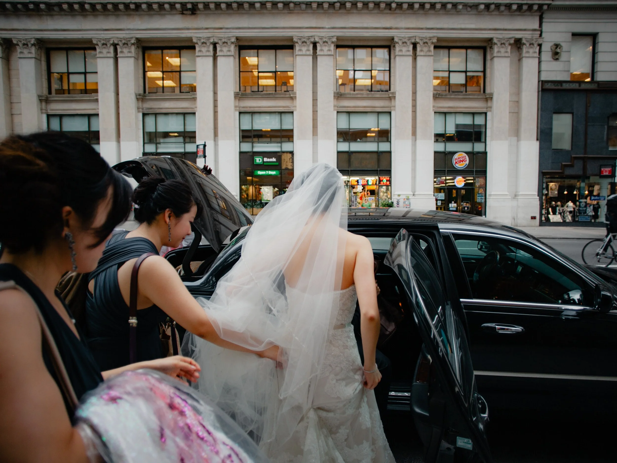 A bride in a wedding dress and veil is getting into a black car, assisted by women standing nearby, on an urban street with shops and buildings in the background.