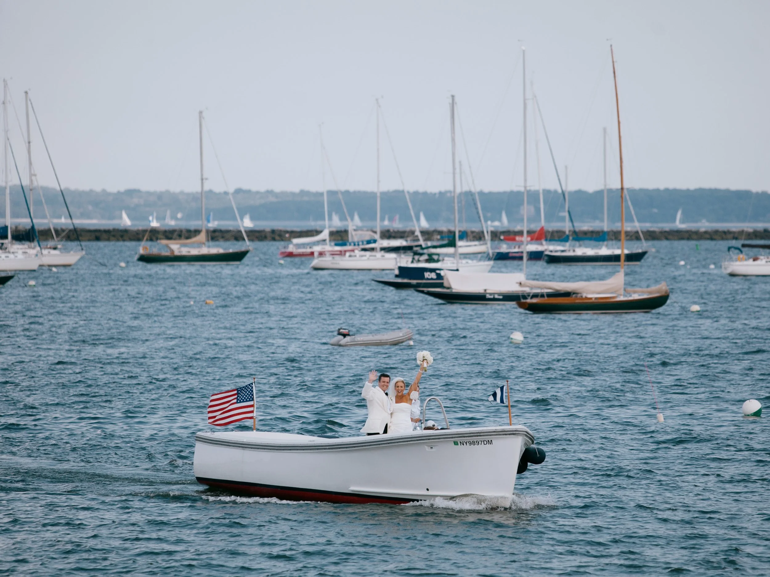 A newlywed couple in wedding attire waves on a small boat with an American flag, on a harbor with sailboats in the background.