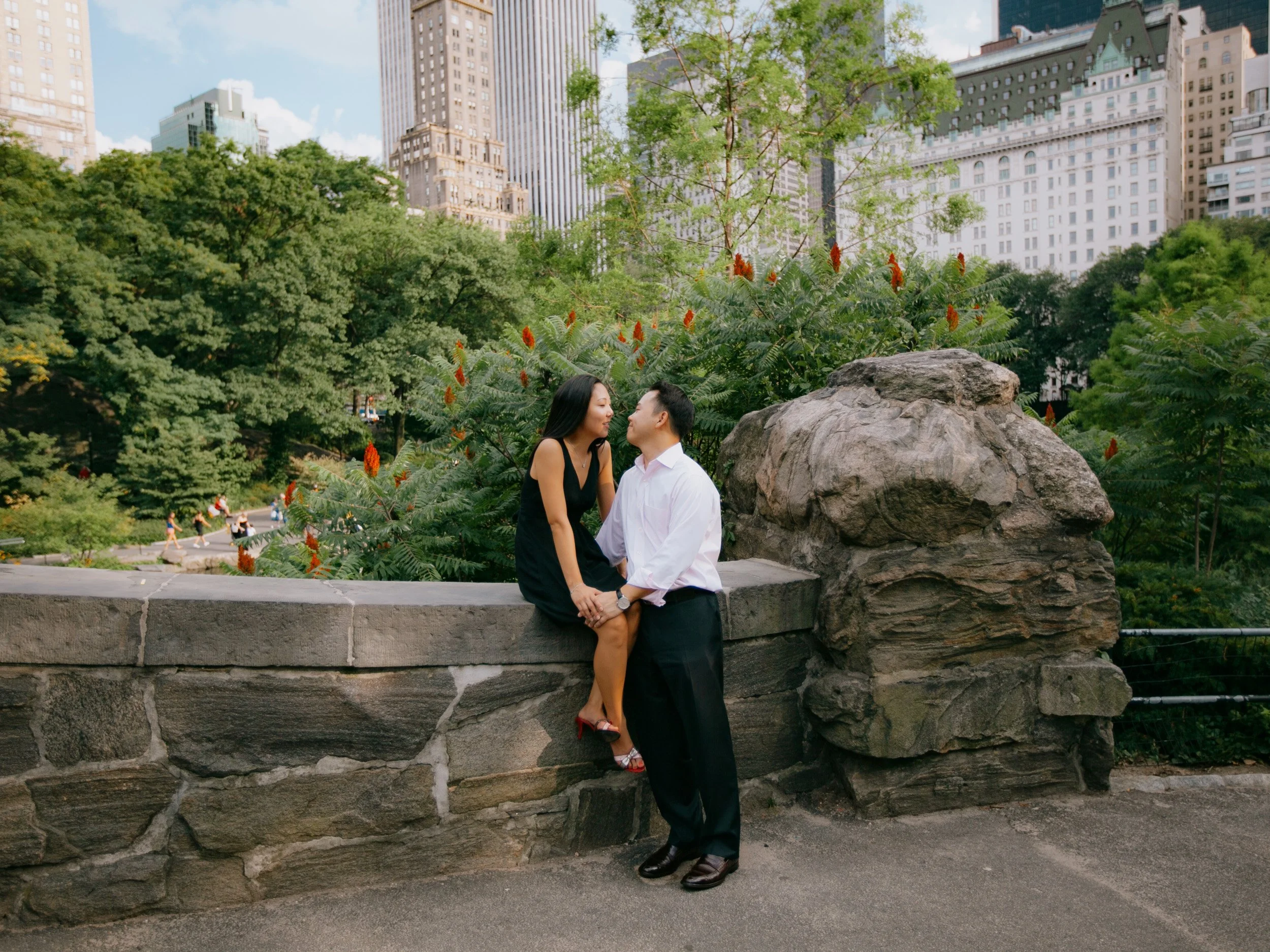 A couple holding hands and leaning in for a kiss at a park in an urban setting, with green trees, colorful flowers, and tall city buildings in the background.