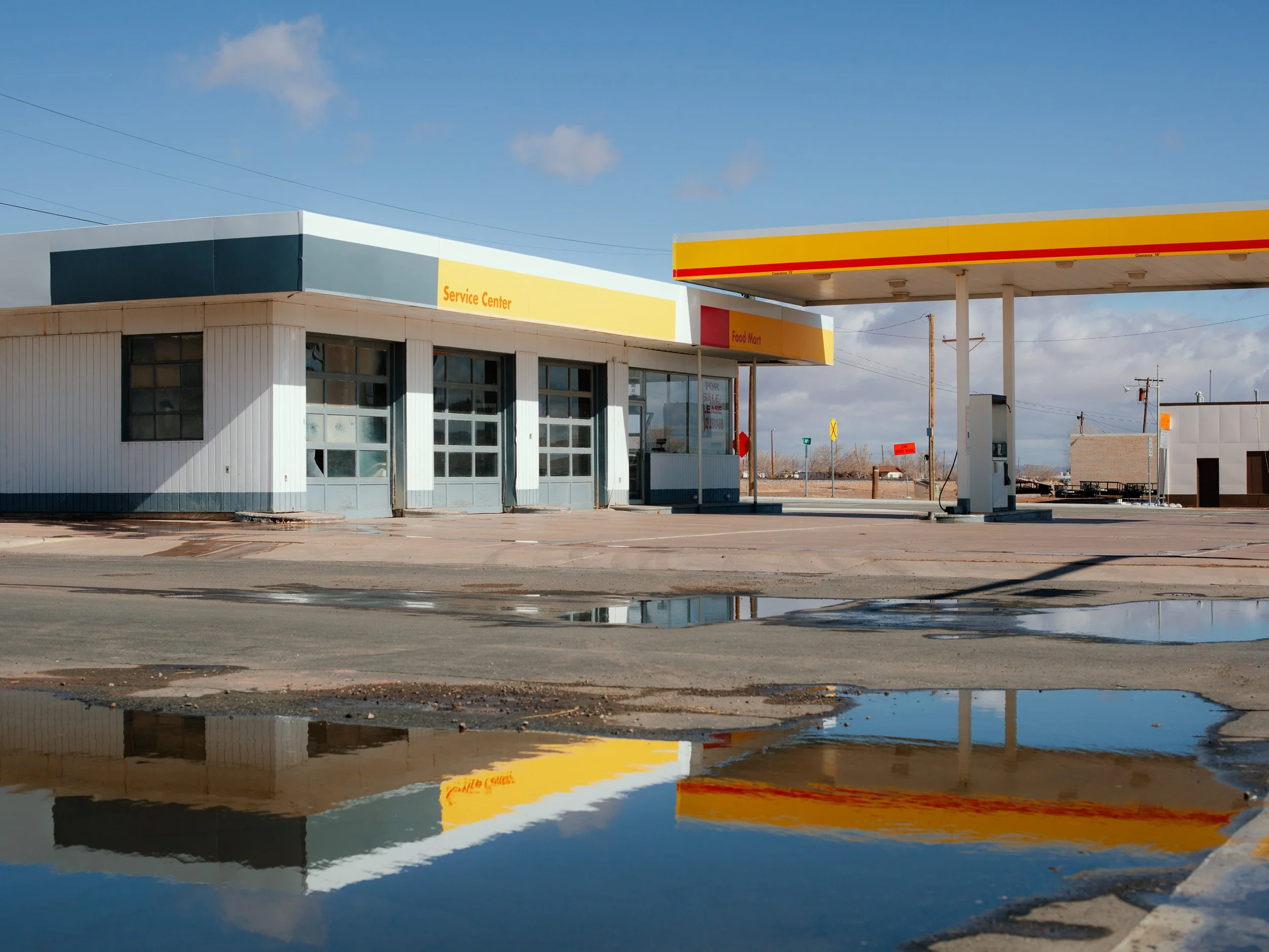 Empty gas station with a Service Center building and a Shell fuel canopy, reflecting in puddles on the ground on a clear day.