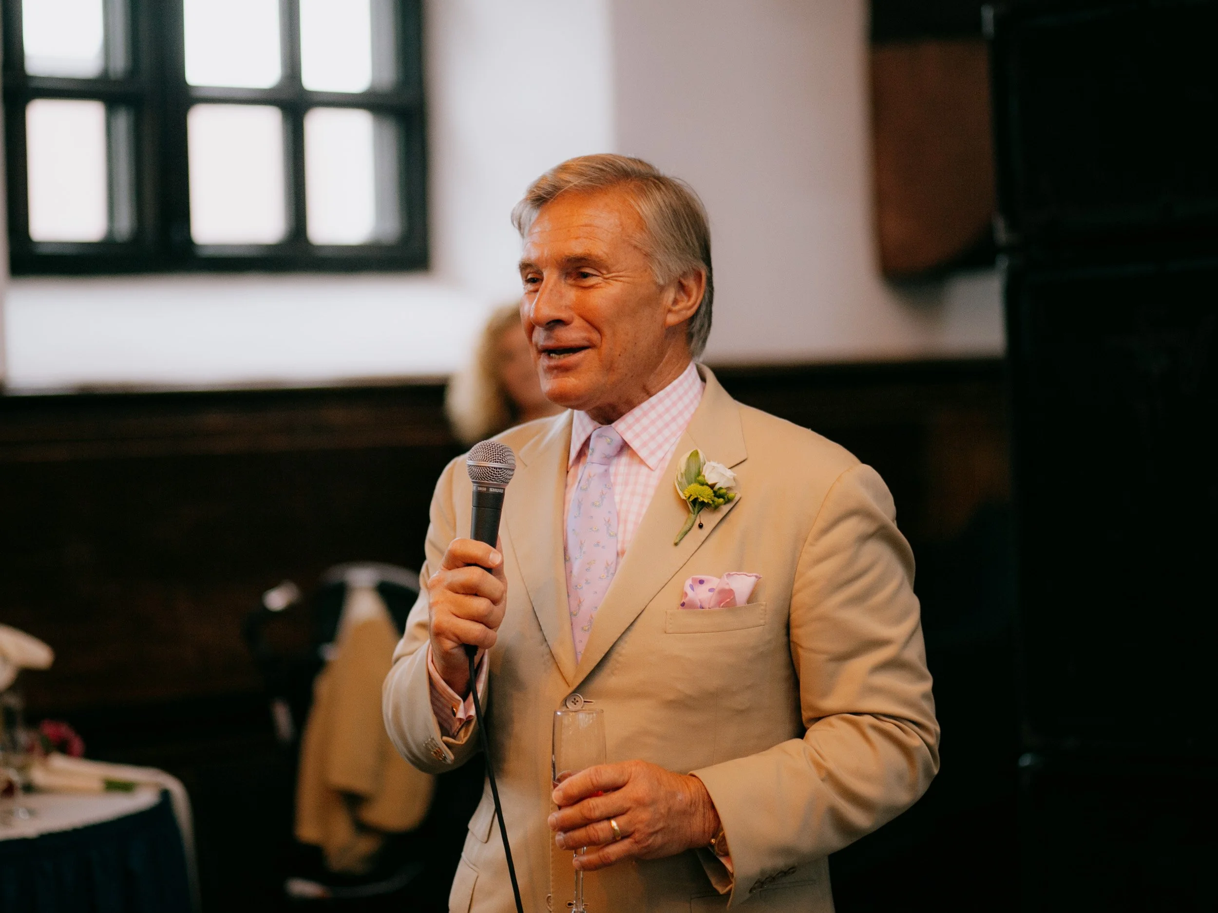 Older man giving a speech at a wedding reception, holding a microphone and a glass of champagne, dressed in a beige suit with a pink checkered shirt and a boutonnière.