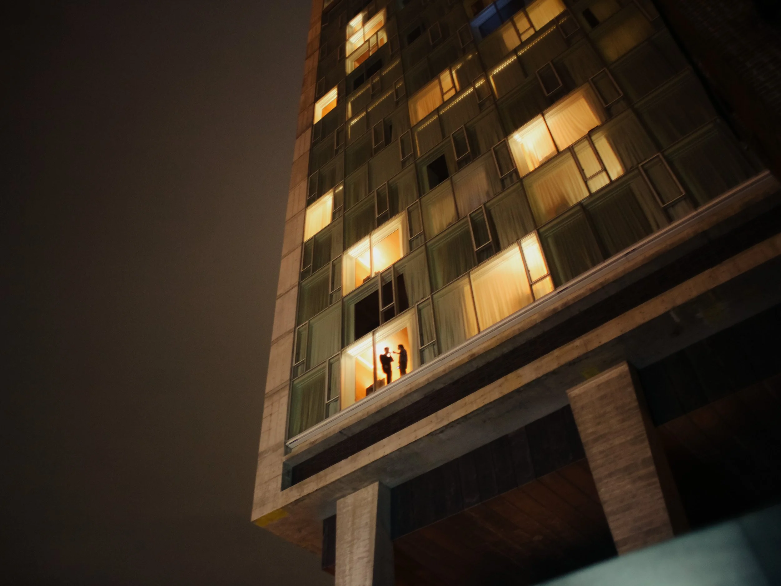 Nighttime view of a tall building with illuminated apartment windows, one of which shows two silhouetted people talking inside.
