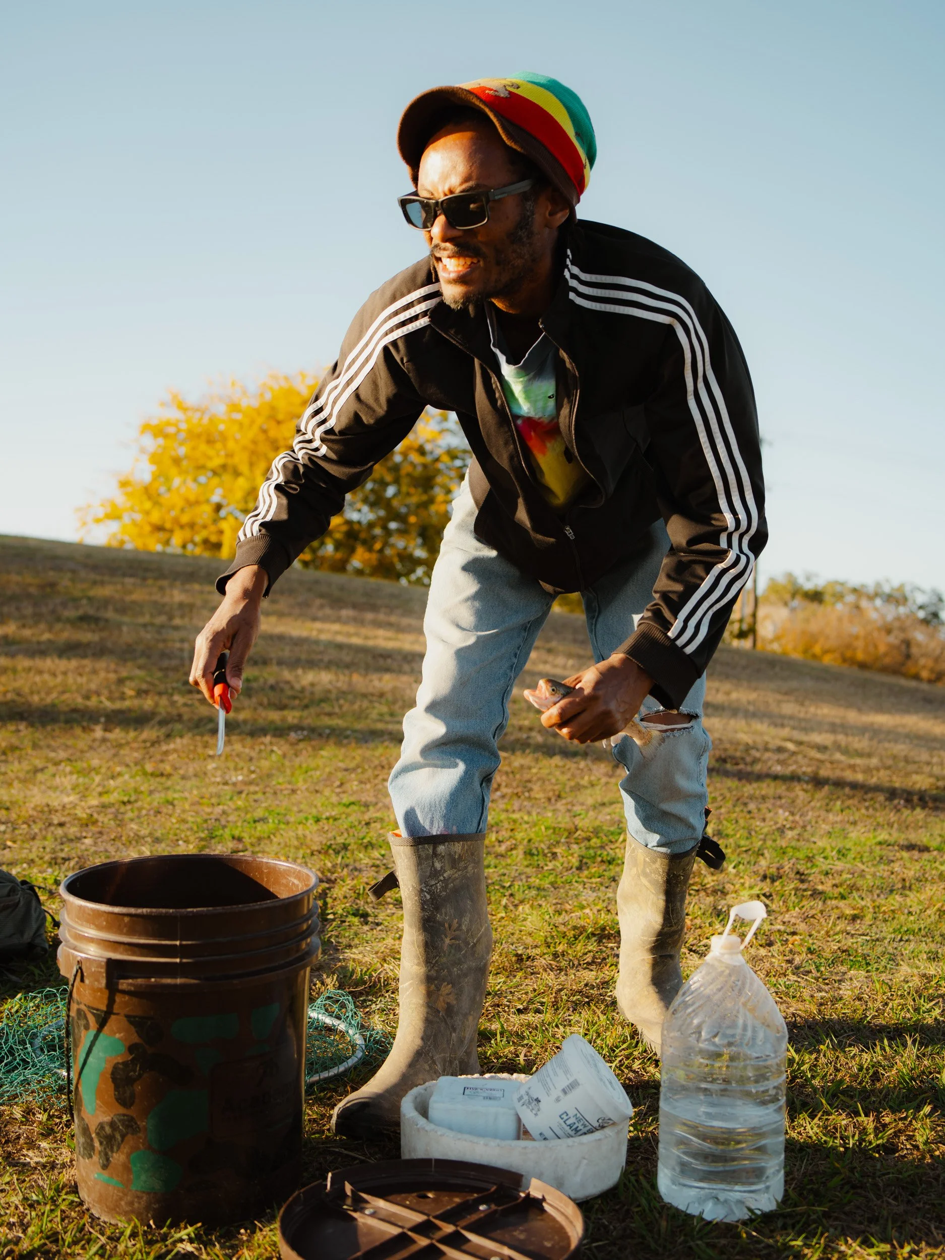 Man wearing sunglasses, a colorful beanie, and rain boots, smoking a pipe outdoors during daytime, with a grassy field and yellow-leaved tree and clear blue sky in the background.