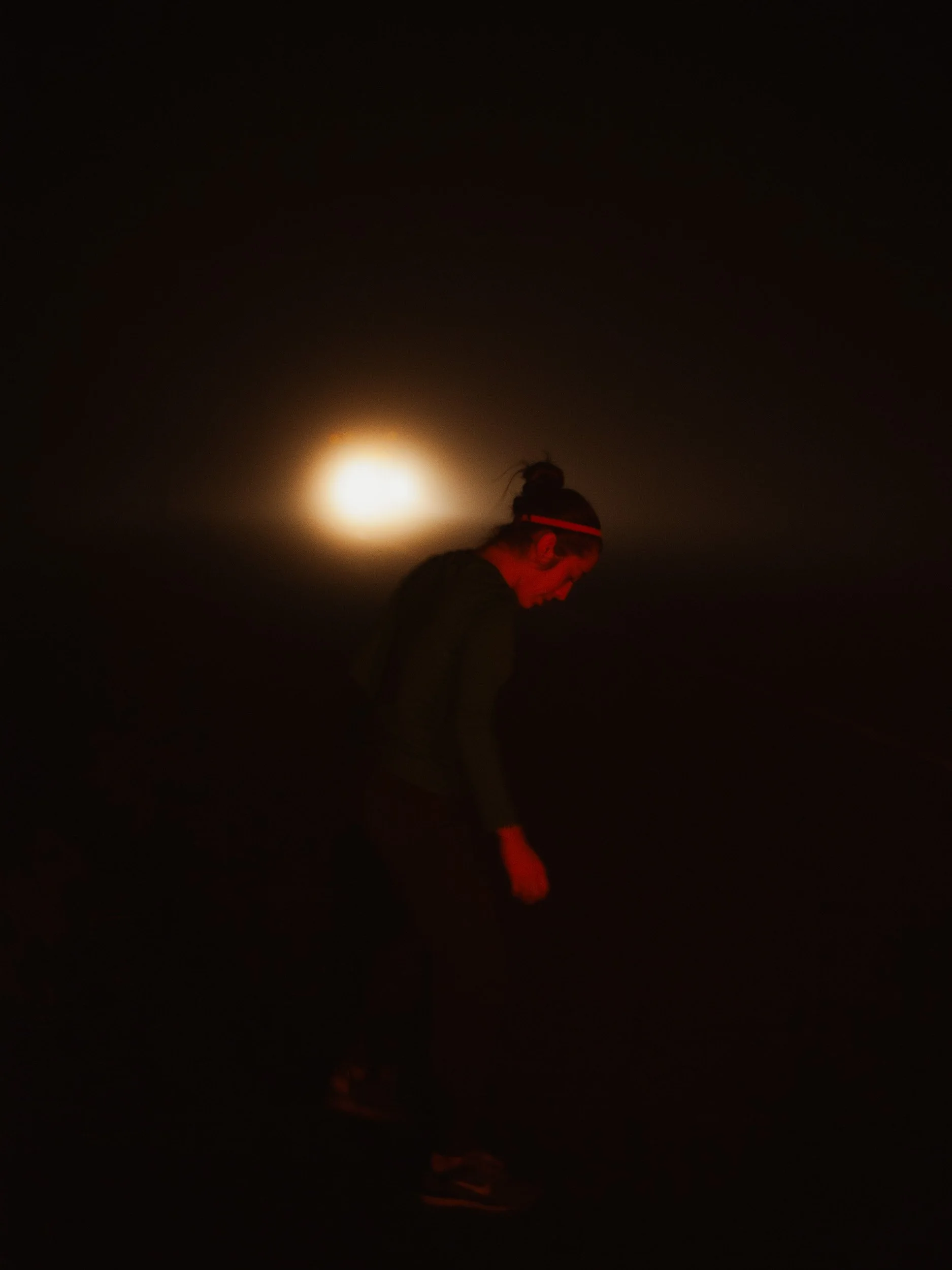 A woman in a dark setting, lit by a bright light behind her, wearing a headband and looking down.